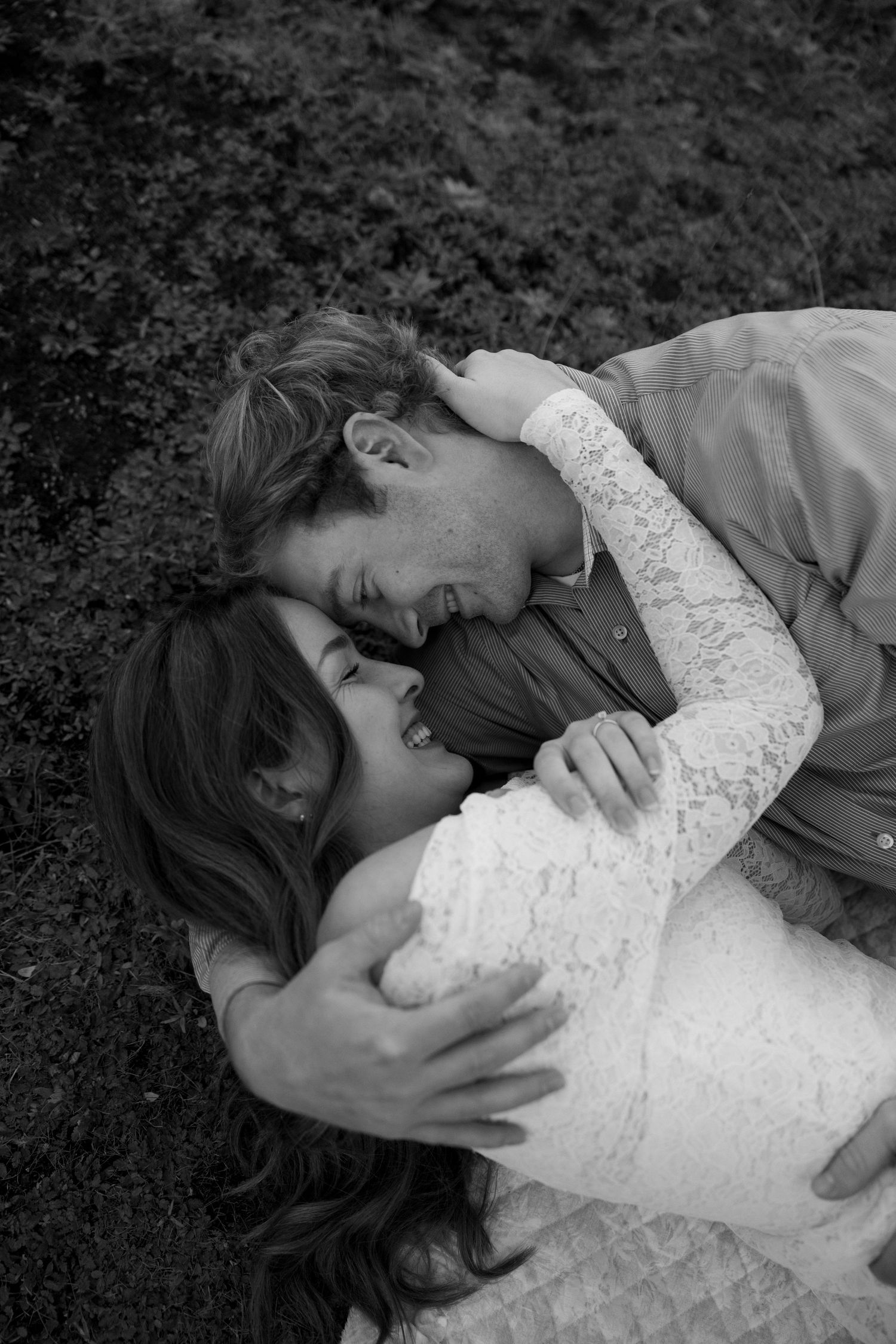 Black and white overhead photo of bride and groom lying on grass, laughing and embracing tenderly.