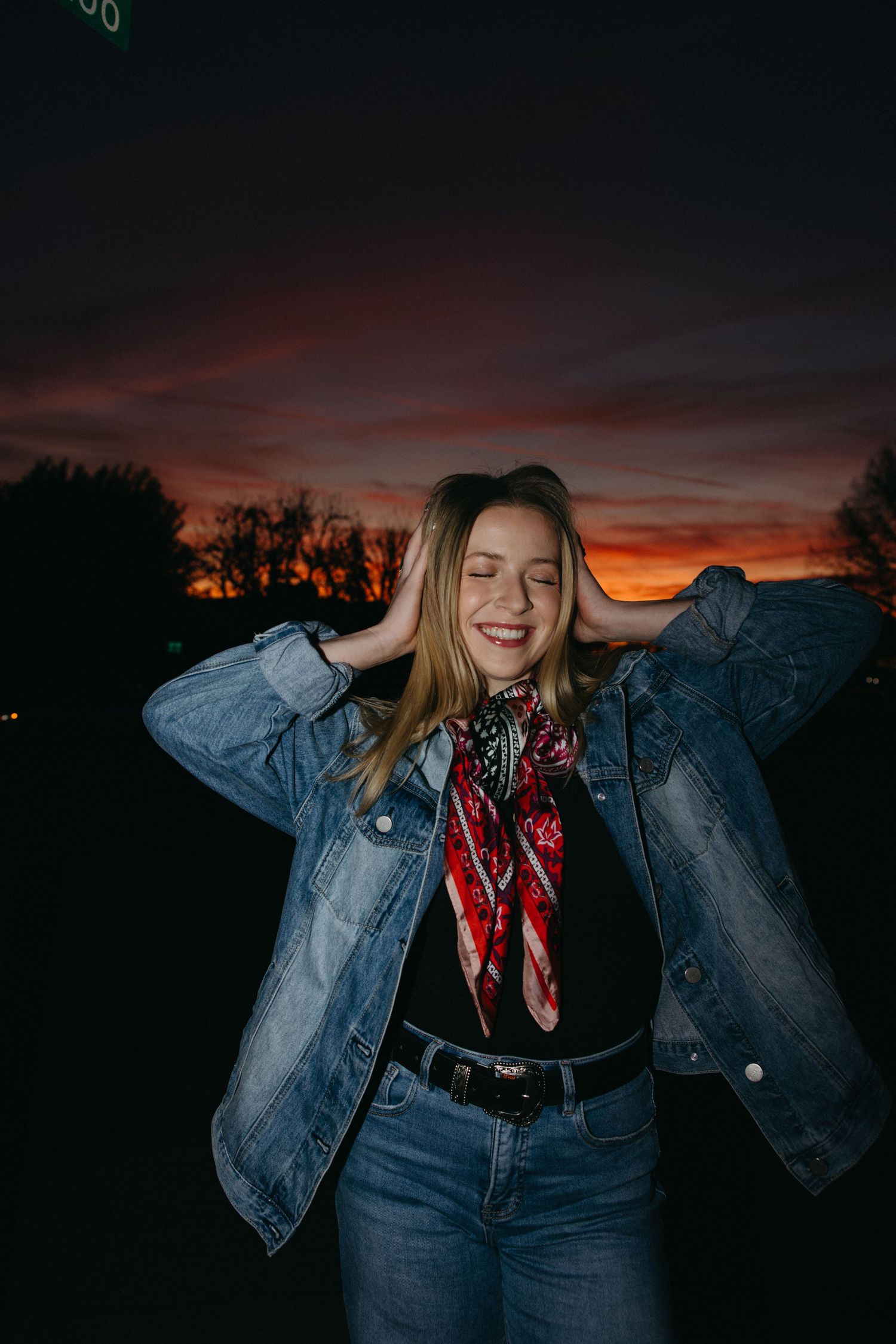 Smiling blonde teen in denim jacket and red scarf poses with hands in hair against a vivid sunset sky.