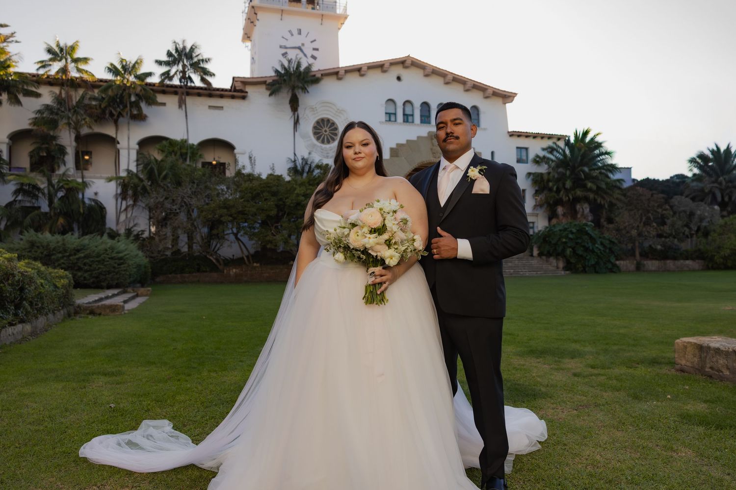Bride and groom posing in front of a historic white building with a clock tower at sunset.