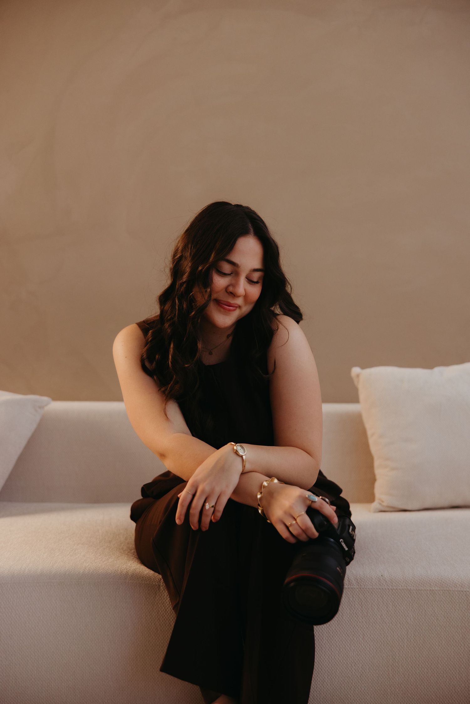 Elegant woman in black sleeveless dress sitting on white sofa looking down at her bracelet.