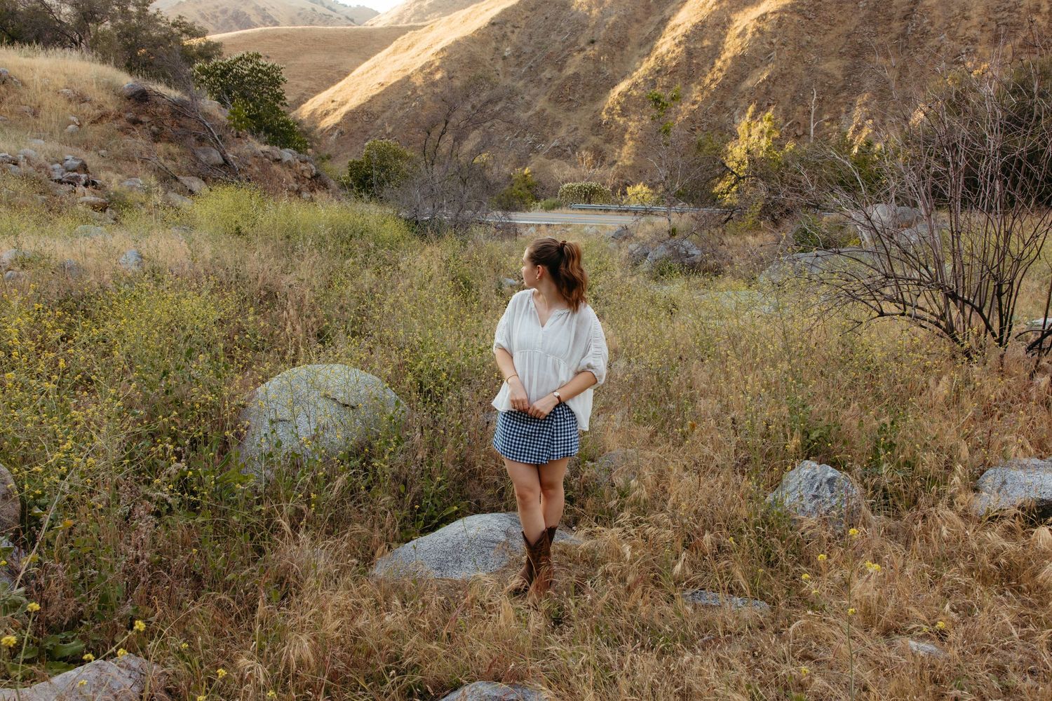Young woman in white top and denim shorts with cowboy boots walking through rocky mountain meadow.