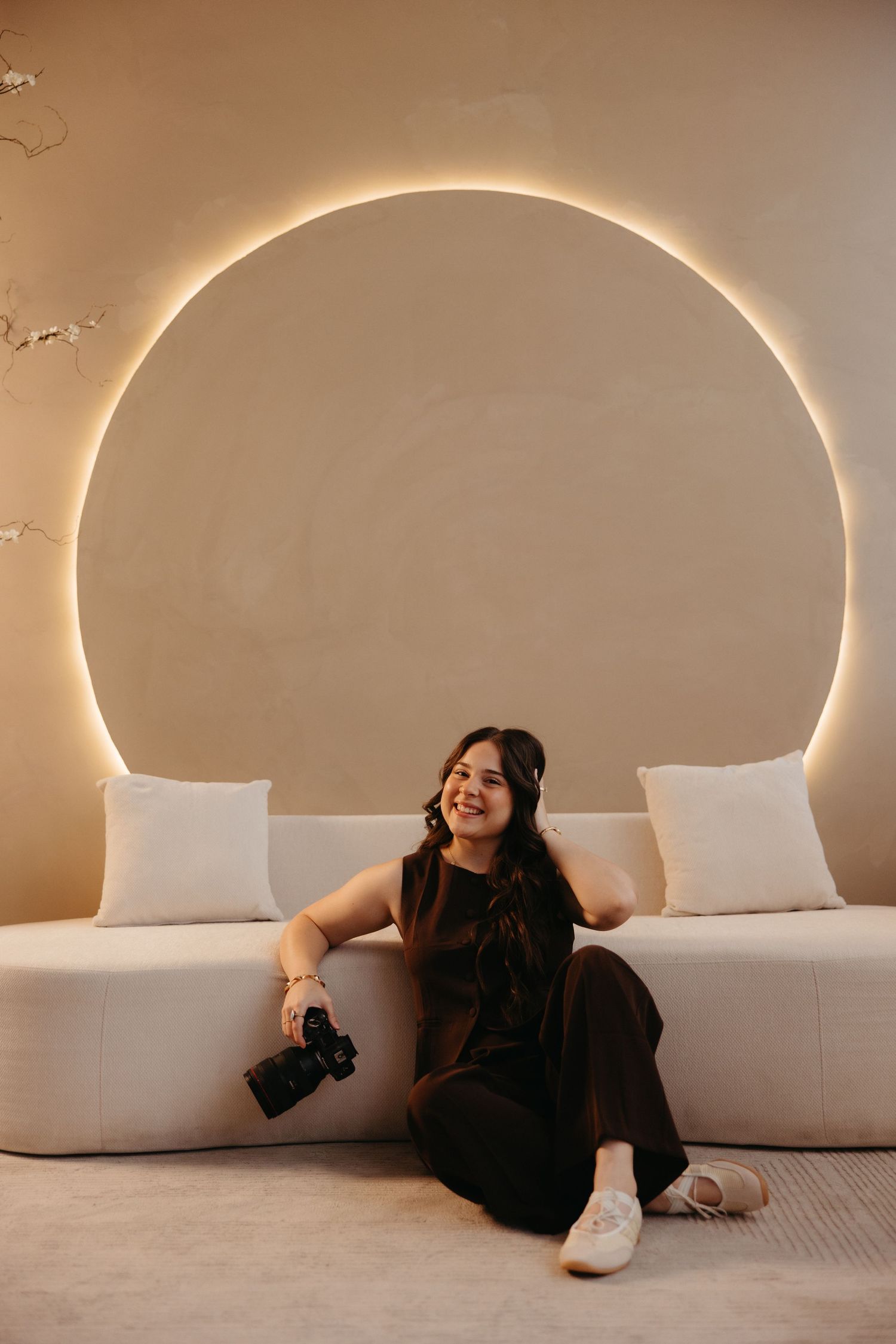 Smiling female photographer sitting on white couch holding camera with glowing circular wall art backdrop.