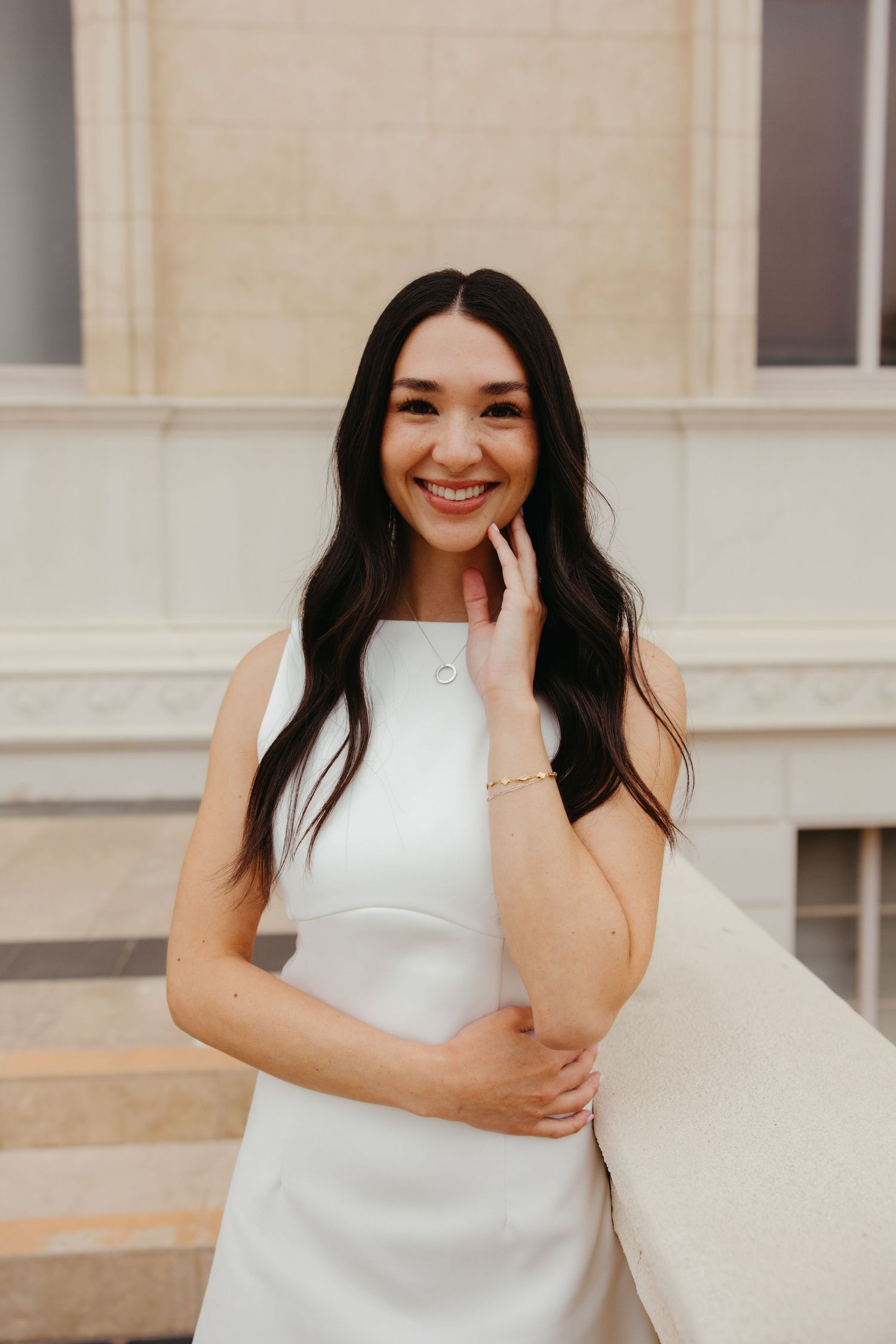 Smiling woman with long dark hair wearing a white sleeveless dress and gold bracelet, posing outdoors near a building.
