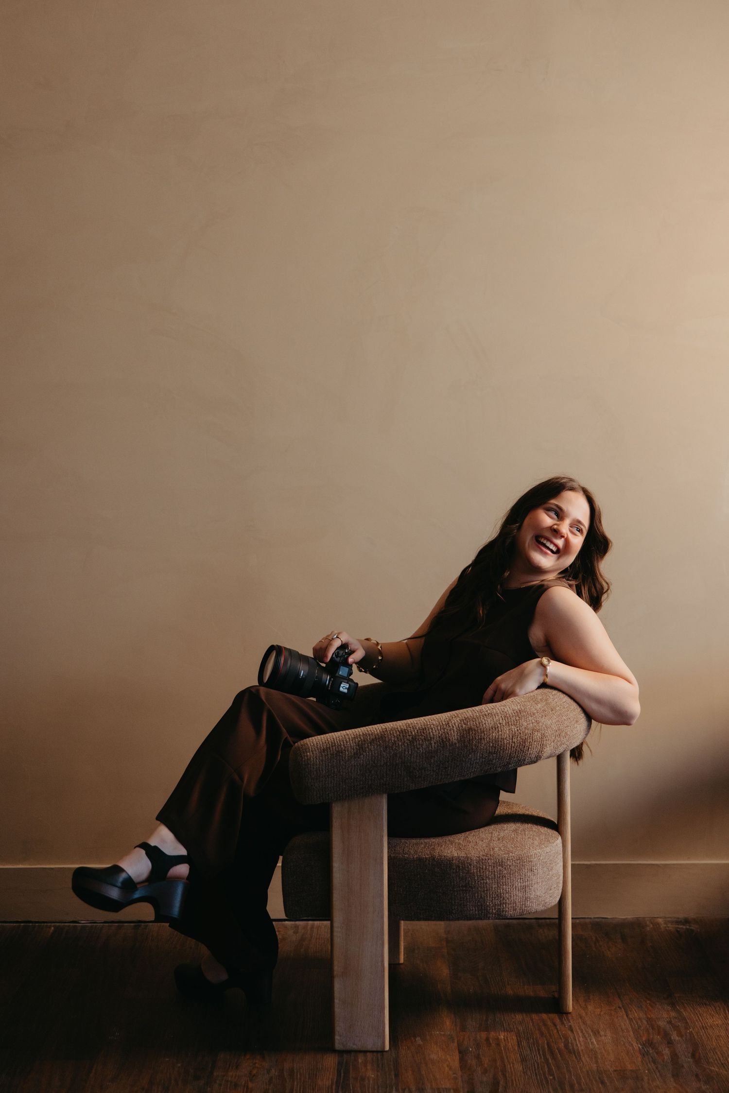 Smiling brunette photographer sitting in a modern chair holding a camera in a warmly lit studio.
