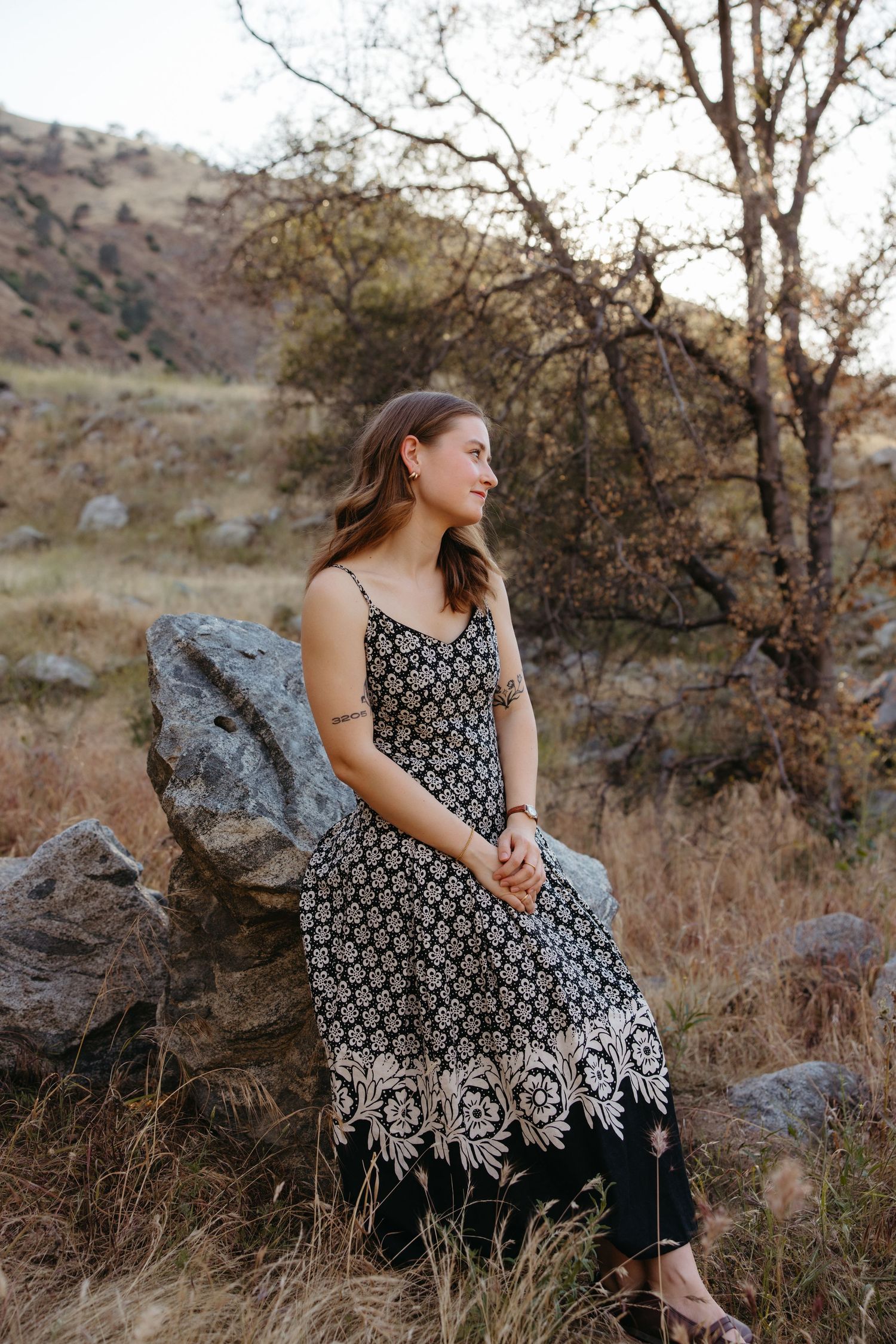 Young woman in black floral maxi dress sitting on rocks in a scenic outdoor field with mountains.