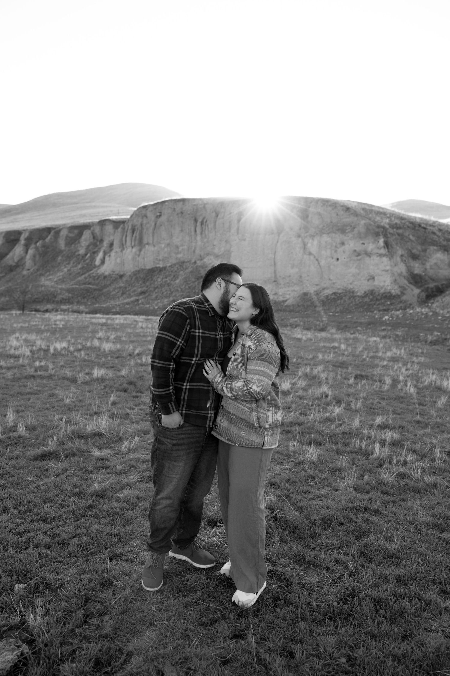 Black and white photo of a couple embracing and kissing in front of a mountain landscape at sunset.