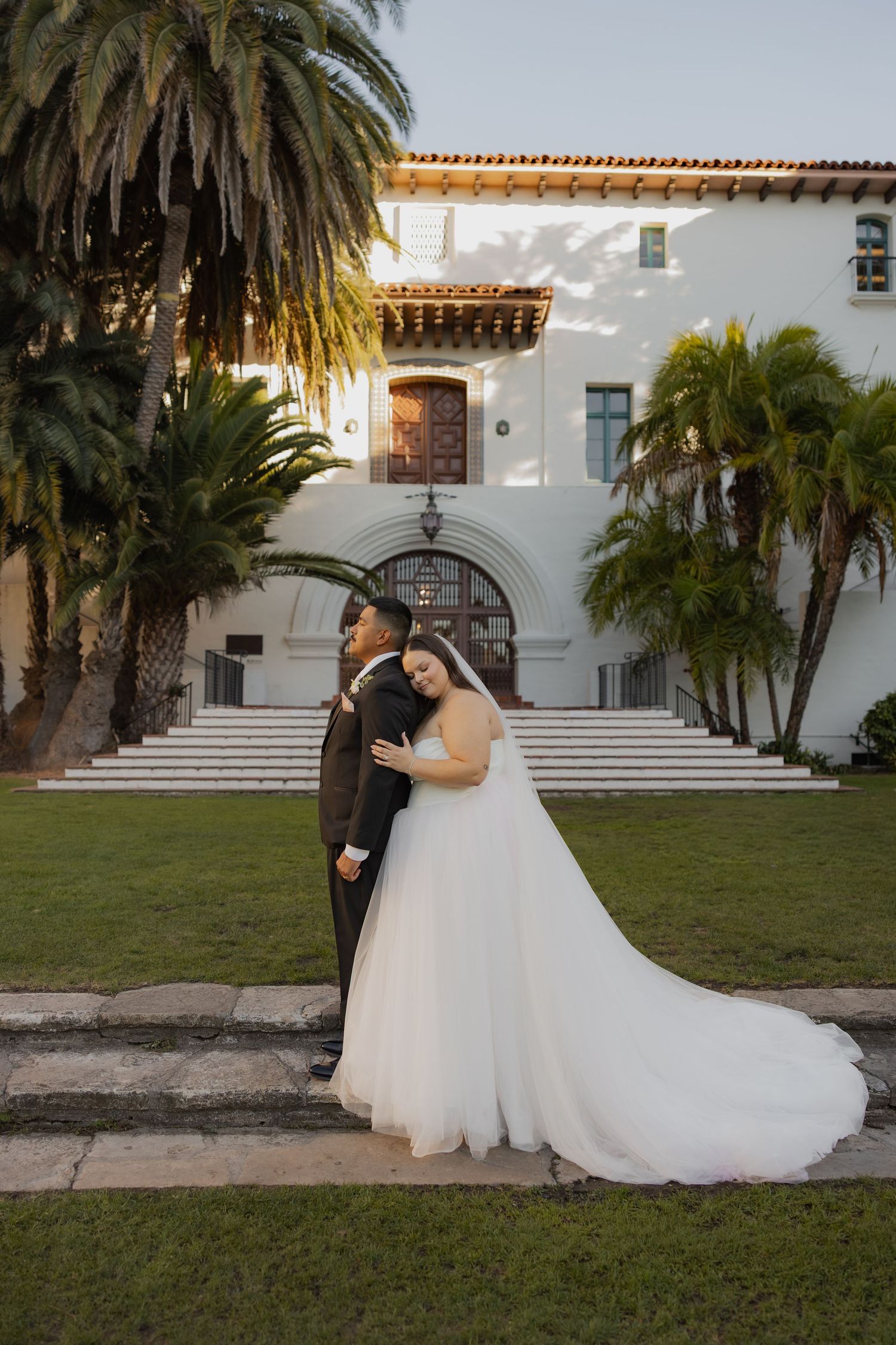 Bride and groom embracing on green lawn before a white Spanish-style wedding venue.