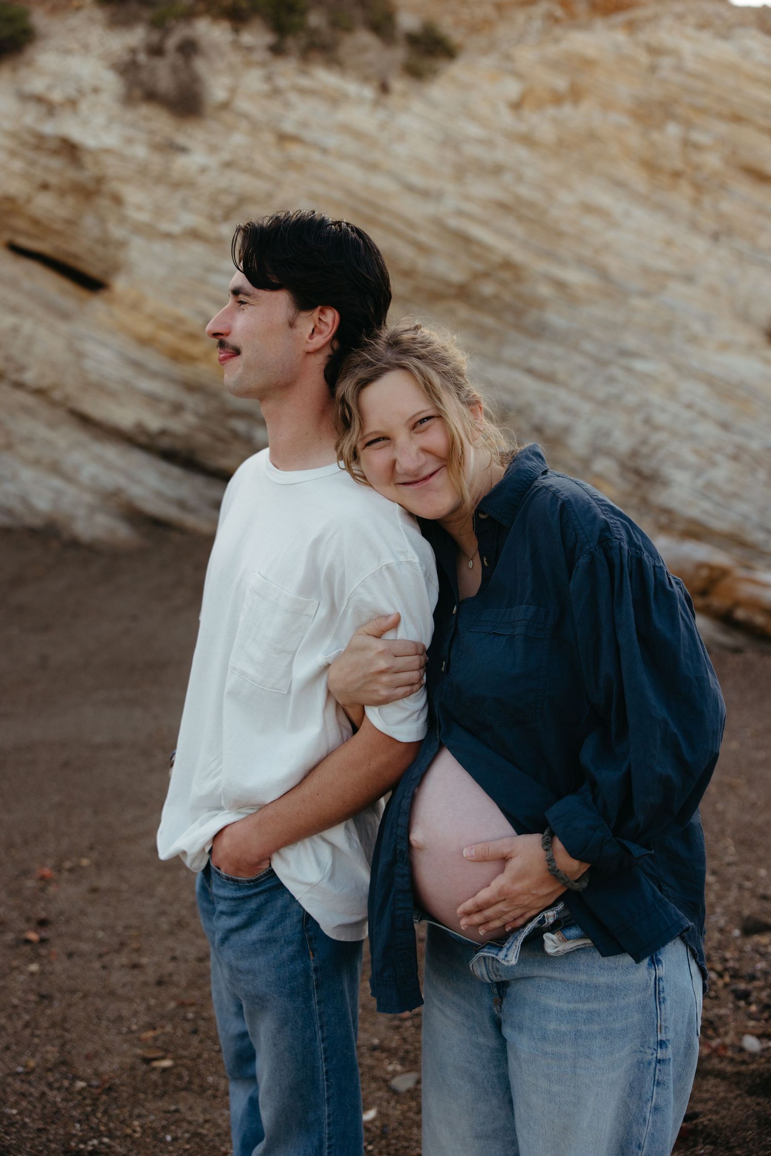 Pregnant woman in denim shirt smiles while embracing partner on a rocky beach at sunset.