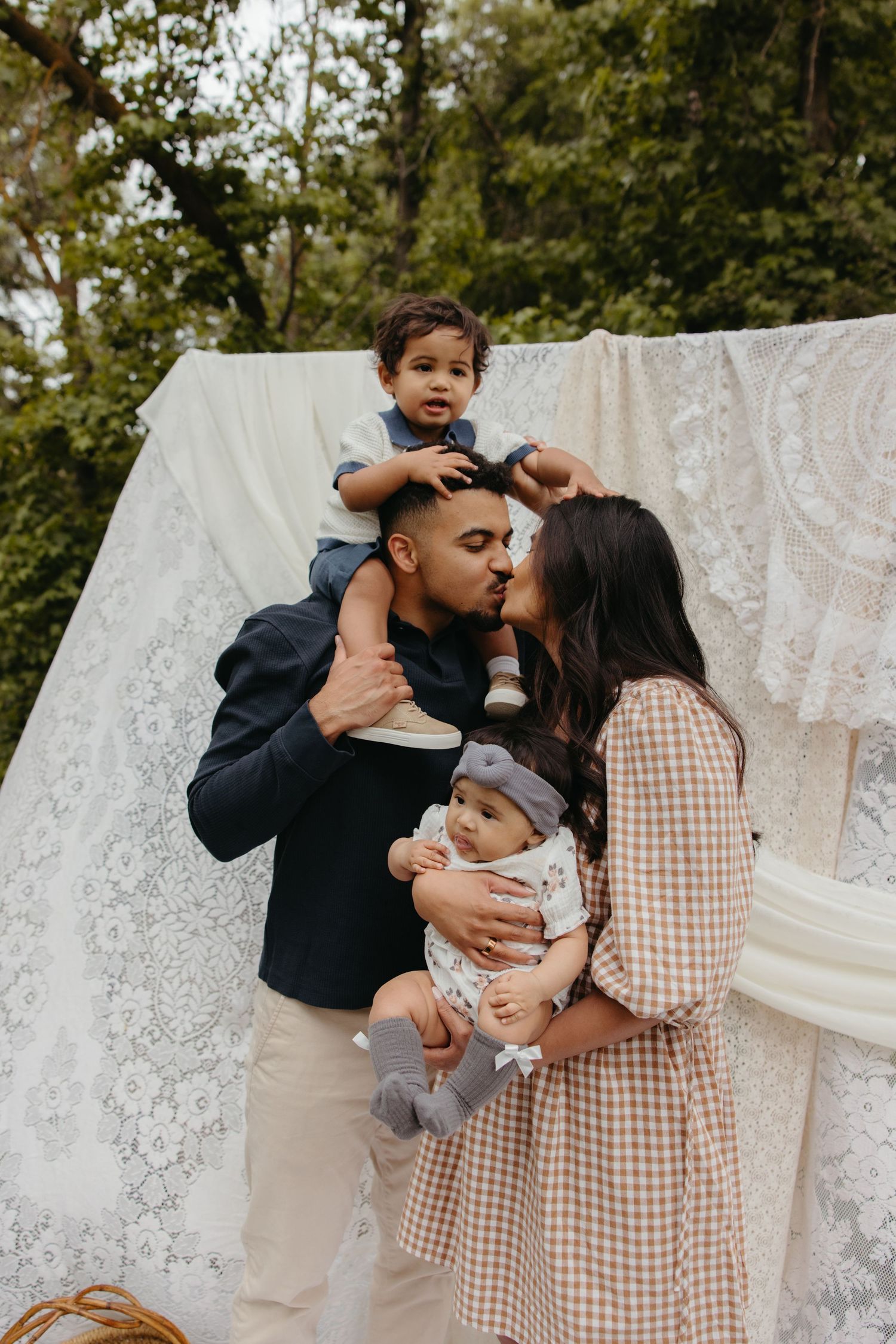 Family of four with two young children kiss and cuddle outdoors before a decorative lace backdrop.
