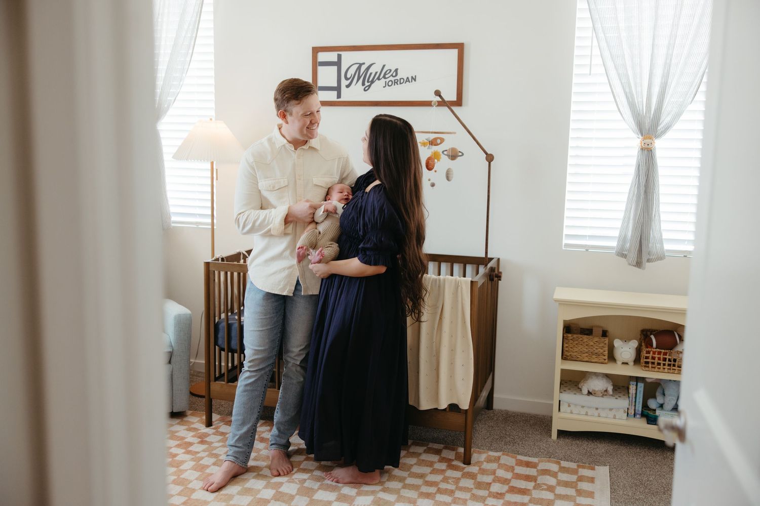 Happy new parents stand together holding their newborn baby in a decorated nursery room.