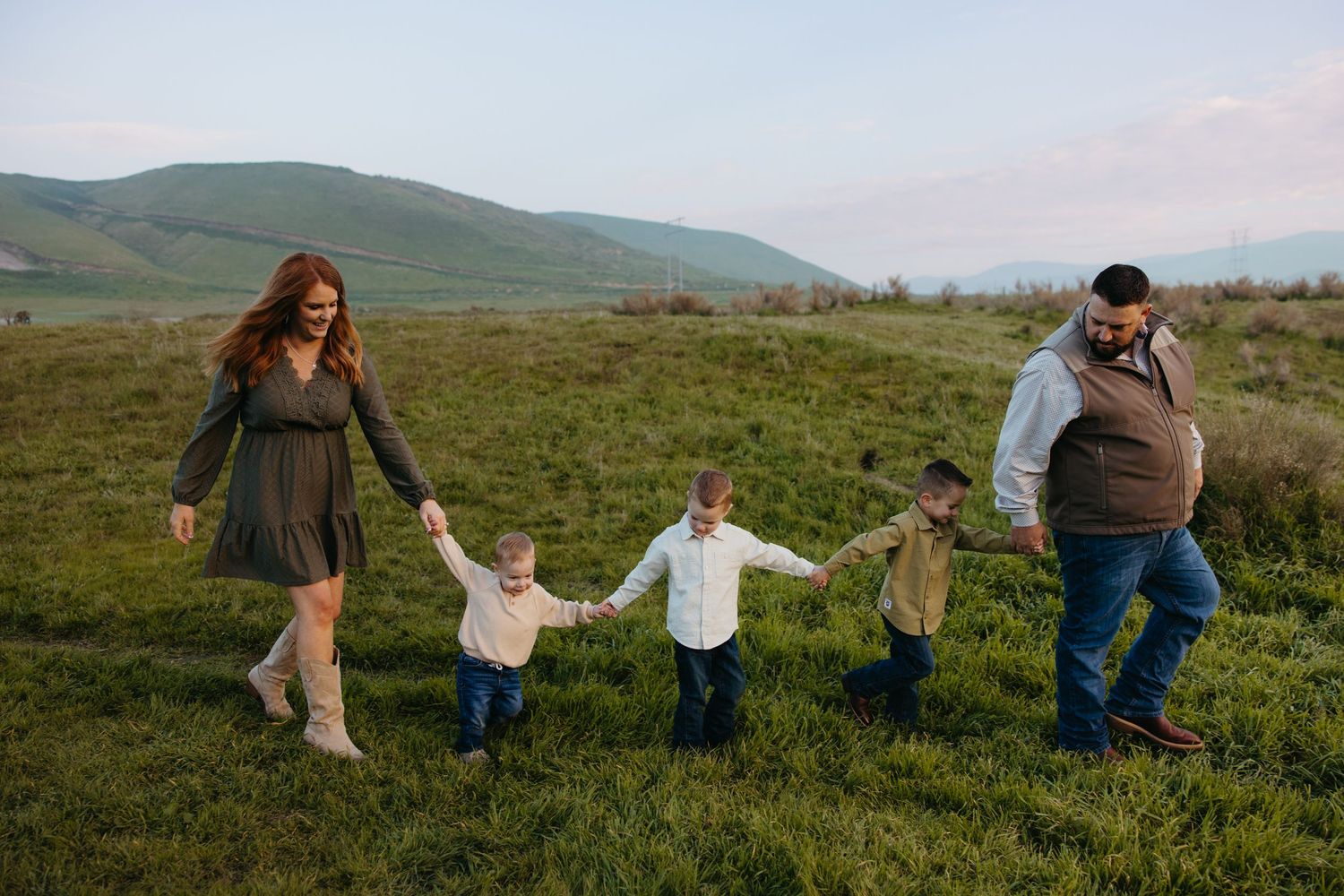 Happy family of five holding hands walking through a lush green field with mountains behind.