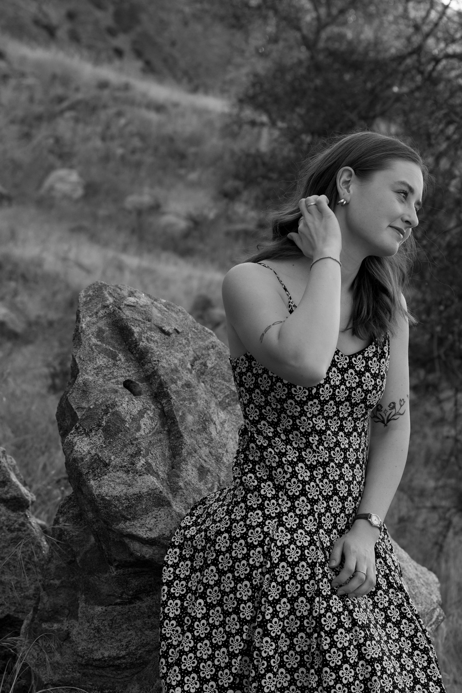 Black and white portrait of young woman in floral dress sitting against a rock in a natural outdoor setting.