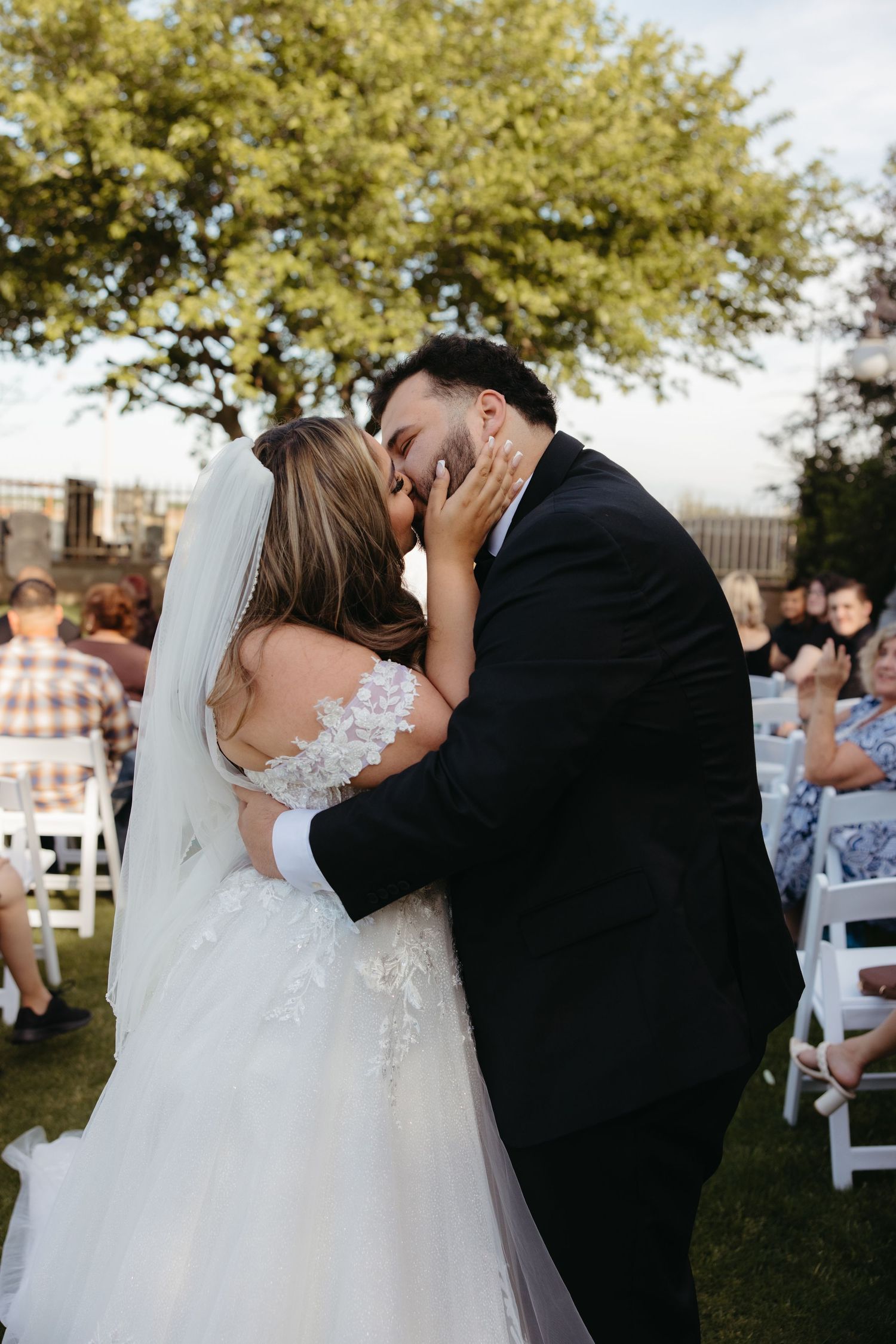 Bride and groom share their first kiss at an outdoor wedding ceremony with guests seated on white chairs.