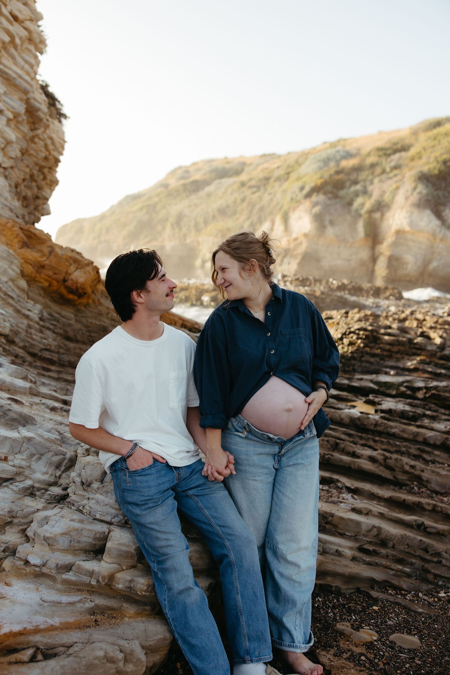 Smiling pregnant couple holds hands while leaning against rocky coastal cliffs in warm sunlight.