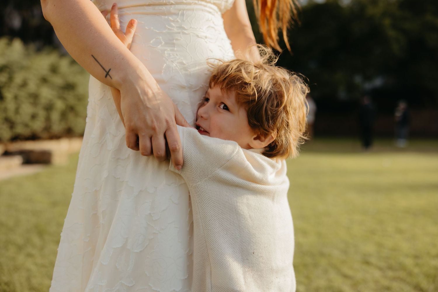 Young curly-haired child hugging mothers white wedding dress hem in sunny outdoor setting.