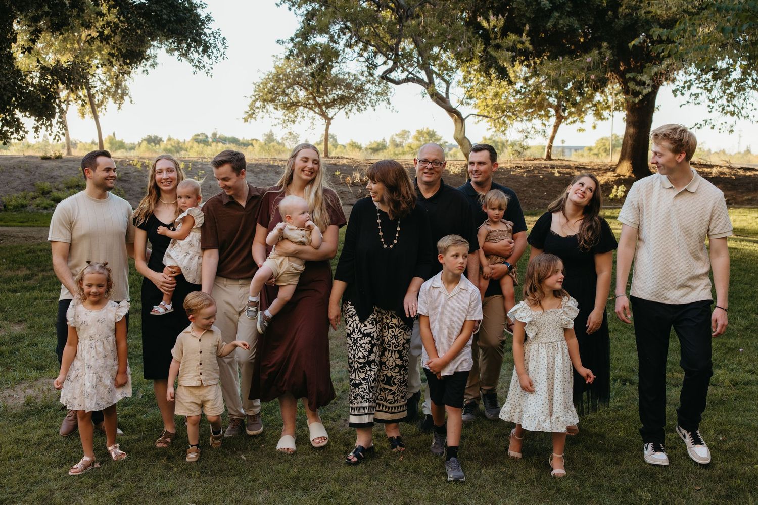 Large multigenerational family posing together outdoors at golden hour under trees on a green lawn.