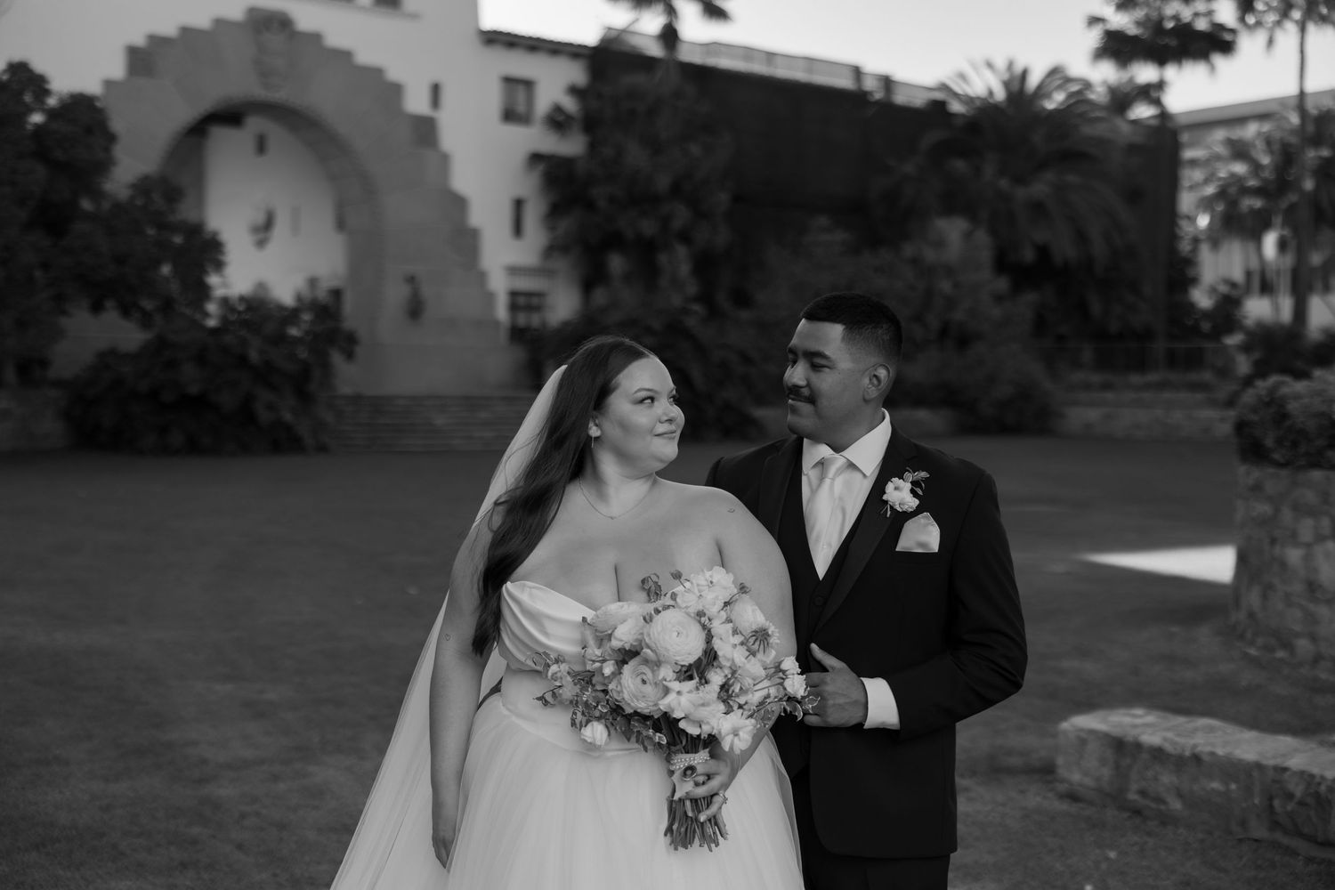 Black and white wedding portrait of bride and groom smiling at each other outside historic Spanish-style venue.