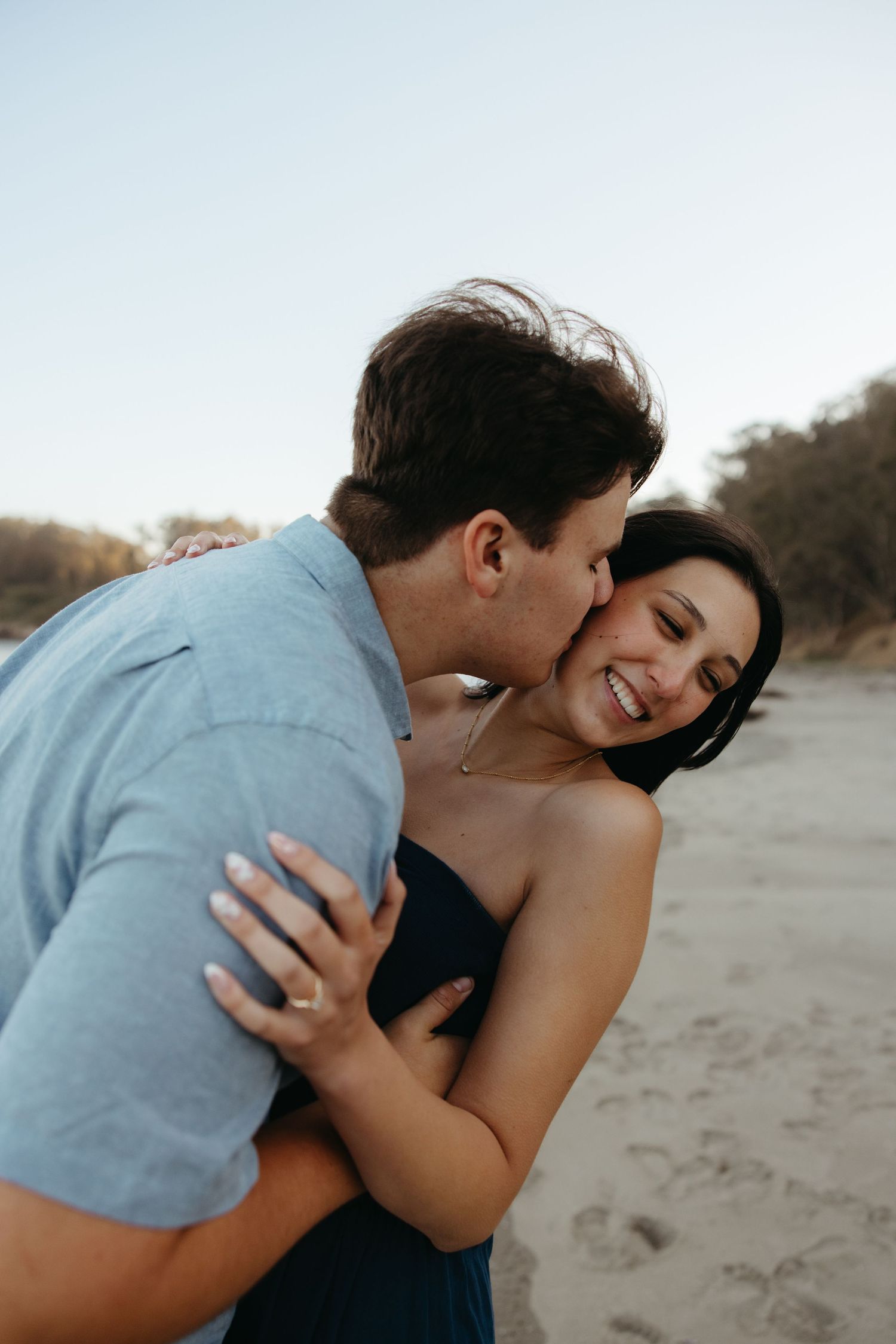 Man kissing smiling womans cheek while embracing her on a sandy beach during golden hour.