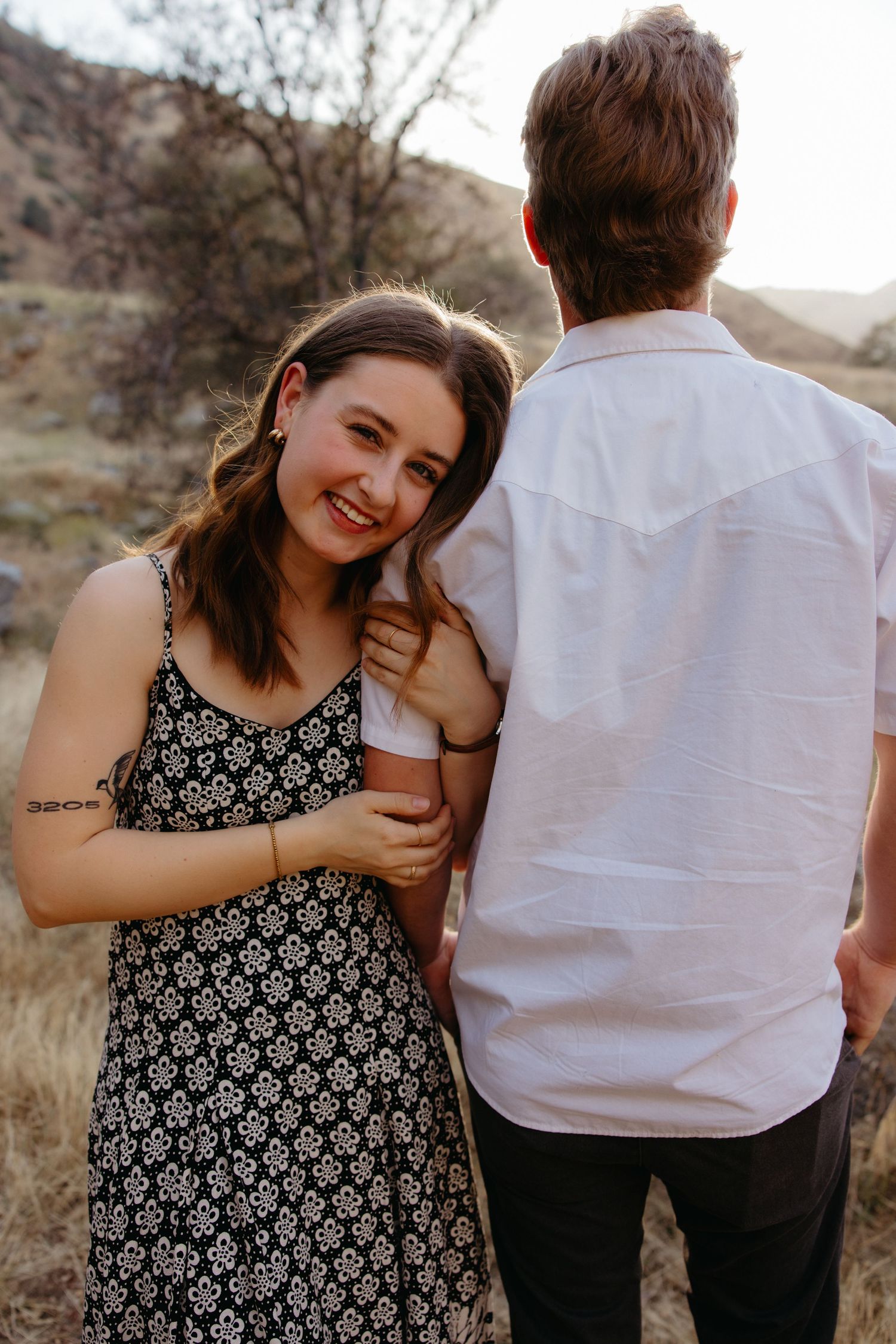A smiling young woman in a floral dress leans on a man in a white shirt outdoors in a dry, grassy landscape.