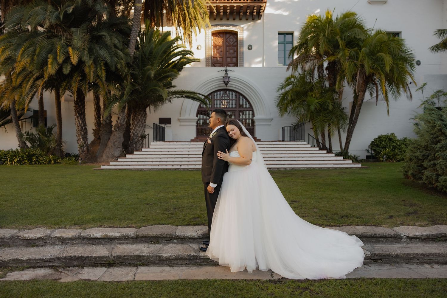 Bride and groom kissing on steps outside a white Spanish colonial wedding venue.