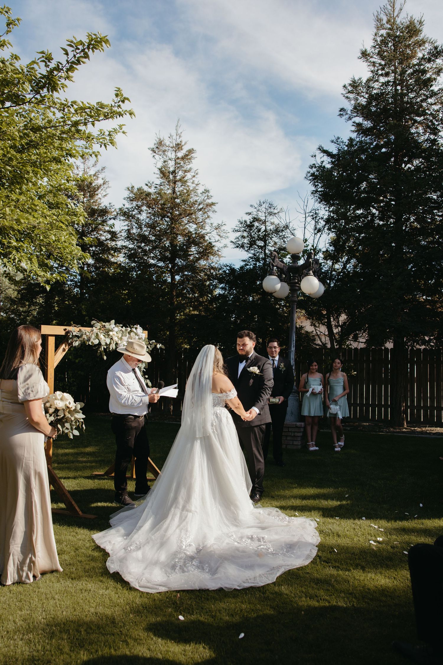 Bride in a flowing lace gown with long veil exchanges vows at an outdoor garden wedding ceremony.