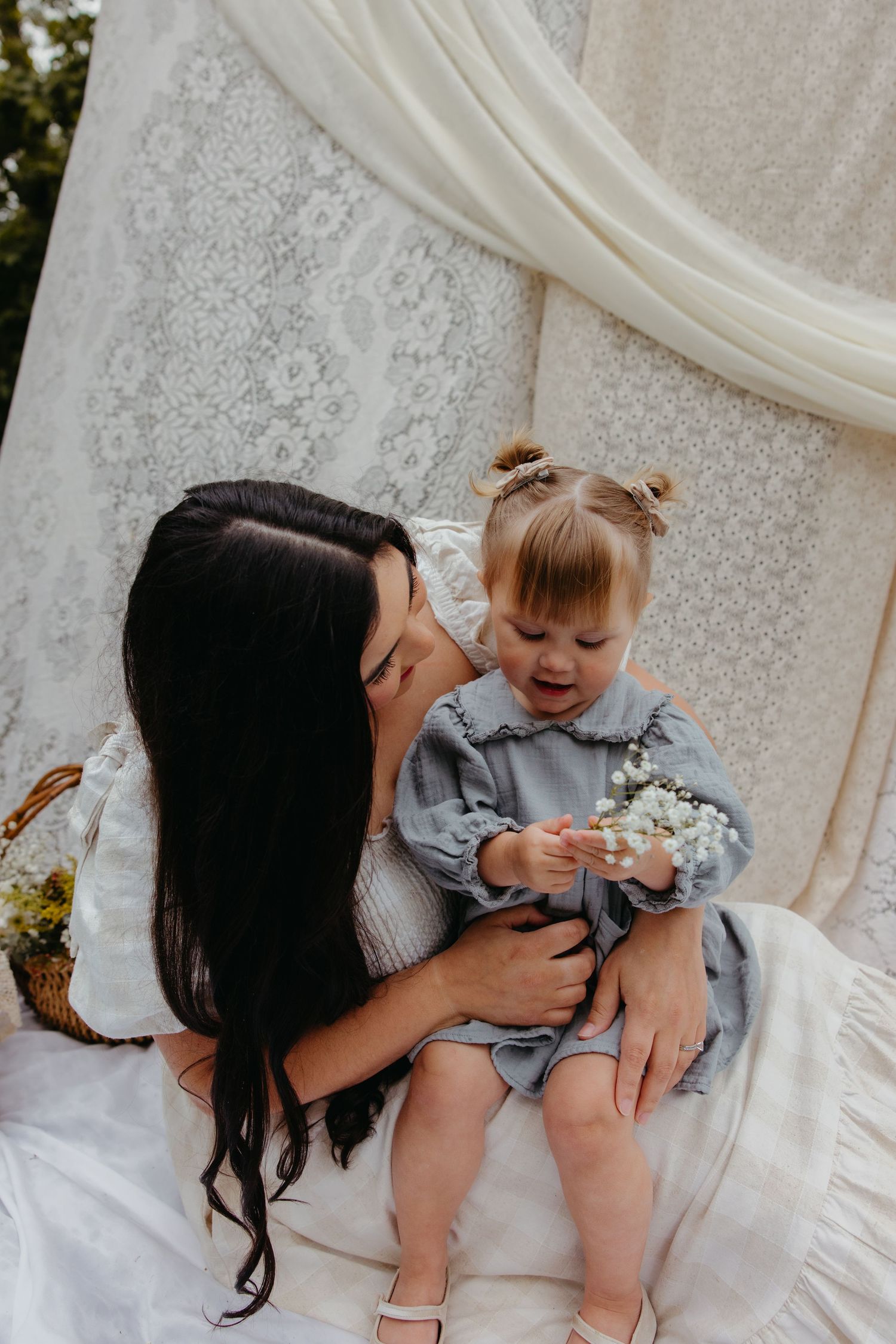 Dark-haired mother tenderly holds smiling toddler girl in blue dress amid white lace backdrop.