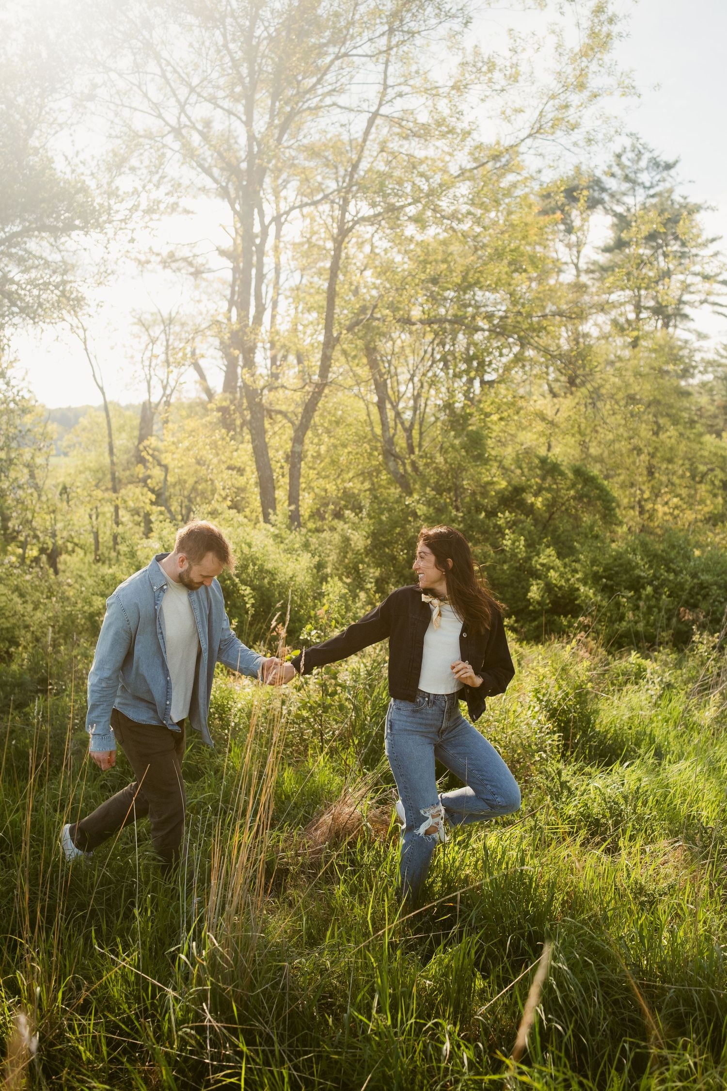Two people enjoying a playful moment together in tall grass during golden hour with sunlit trees in the background.