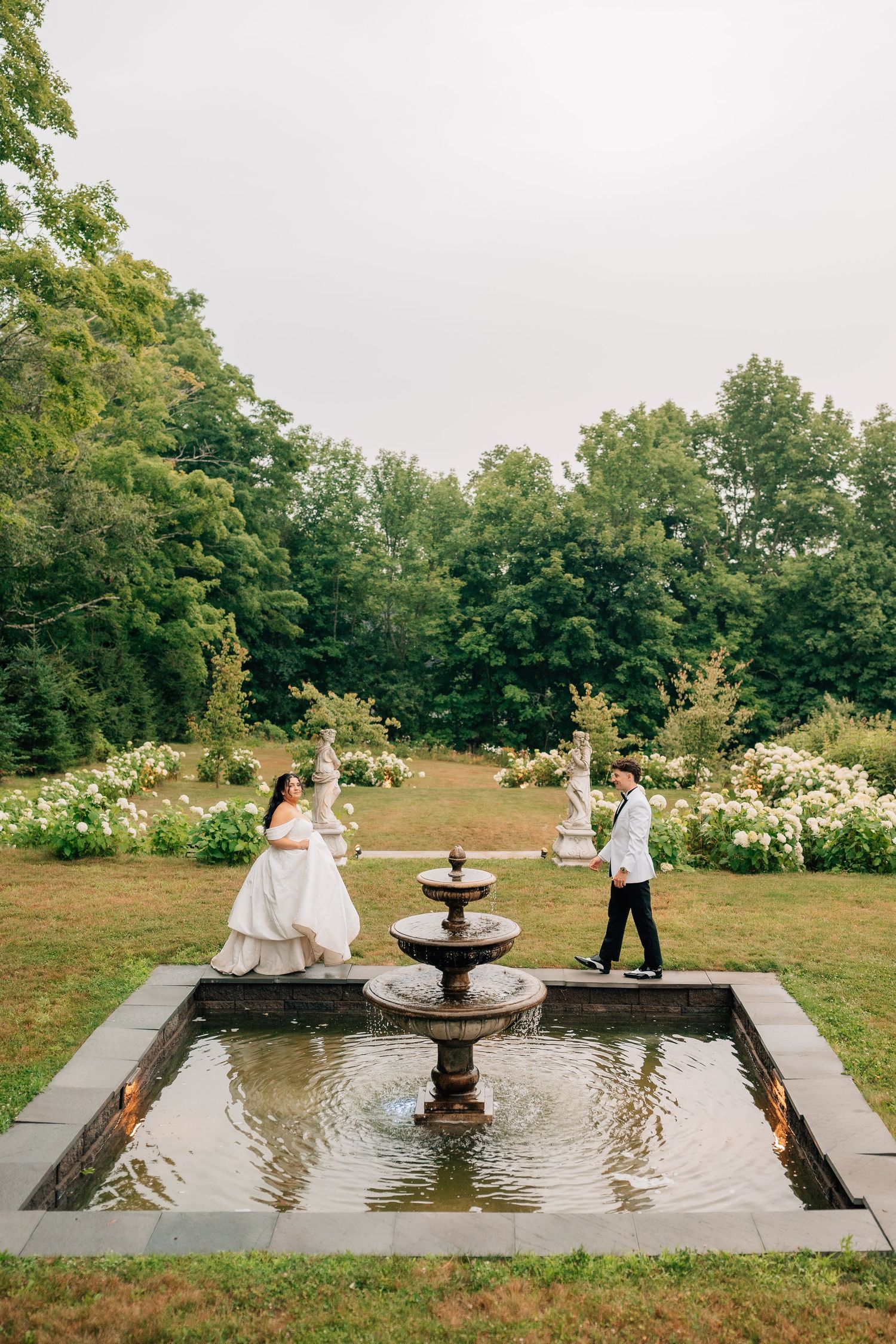Bride and groom standing by a stone fountain in a lush garden with white roses and tall trees at their wedding.