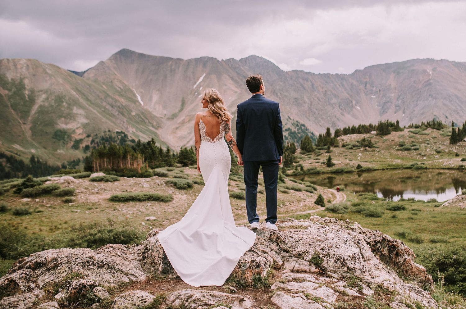 A couple in wedding attire stand on a rocky overlook with majestic mountain peaks and alpine meadows in the background.