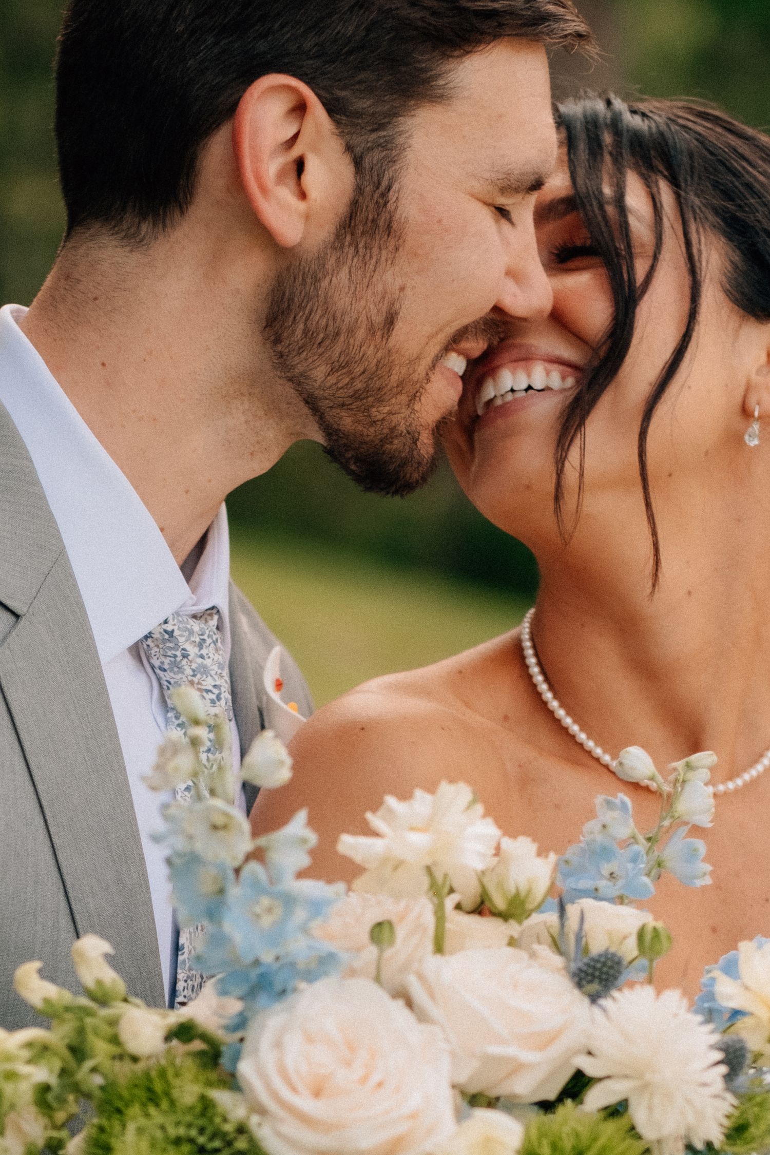 Bride and groom smiling closely together, holding white and blue wedding bouquet with roses and thistle outdoors.