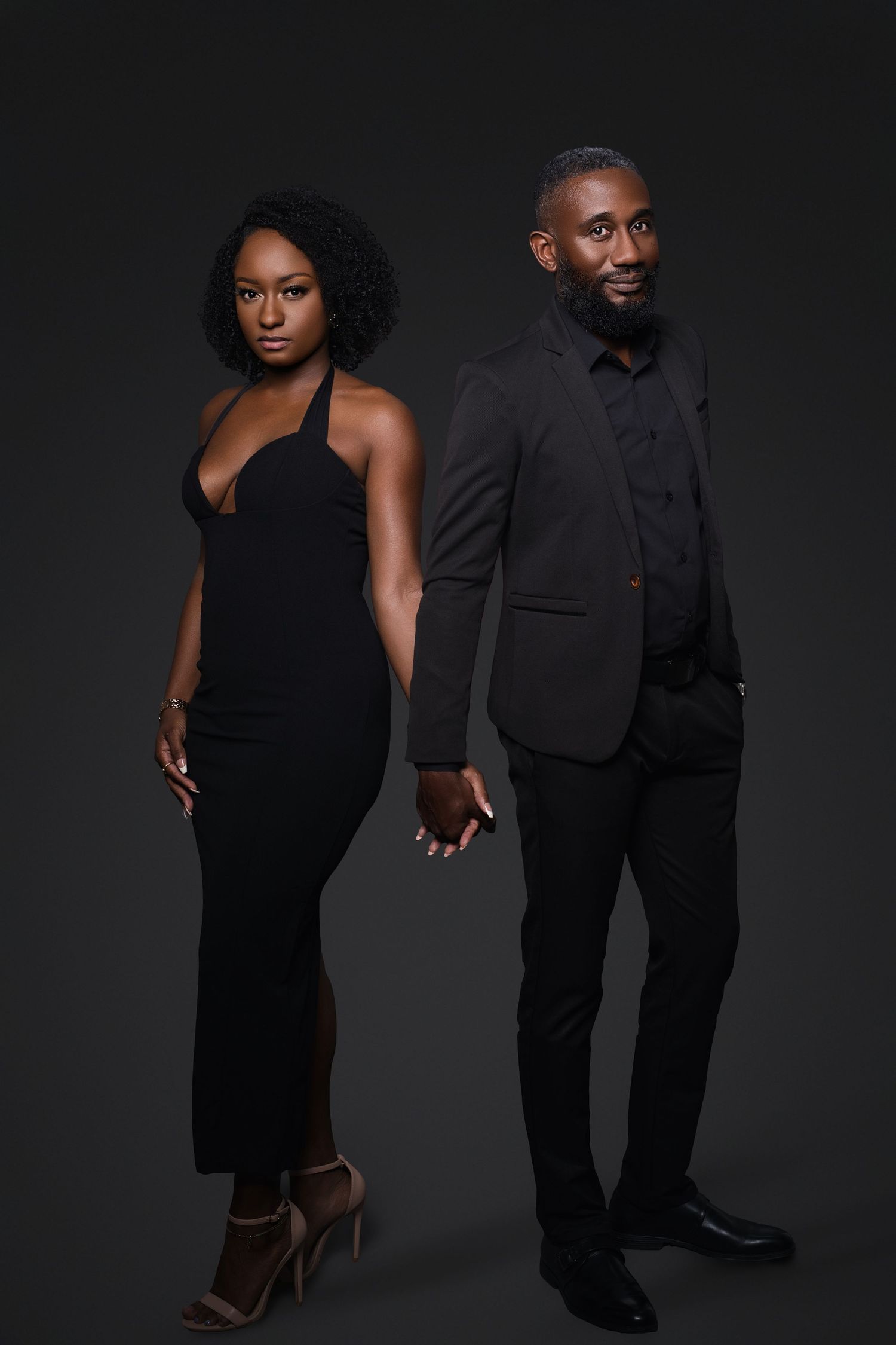 Elegant Black couple in matching black formal attire posing together against a dark studio background.