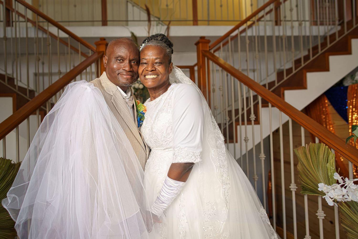 Happy couple in wedding attire smiling together near staircase, bride in white gown and groom in suit with veil.
