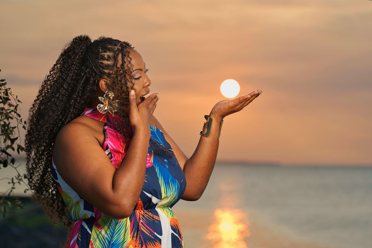 Woman in colorful dress poses at sunset beach, appearing to hold the glowing sun in her outstretched palm.