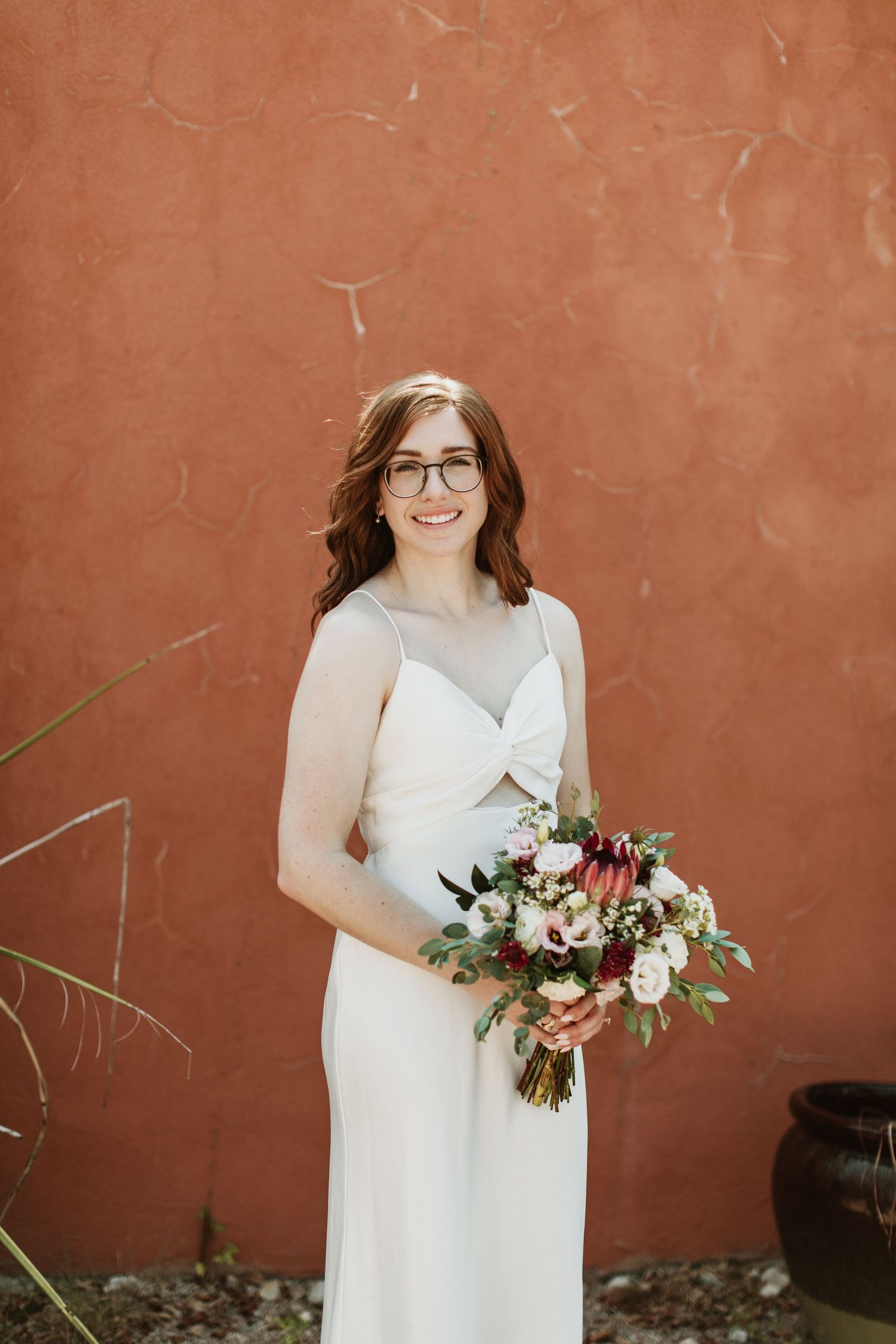 Smiling bride in white dress and glasses holding a burgundy and blush floral bouquet against a terracotta wall.