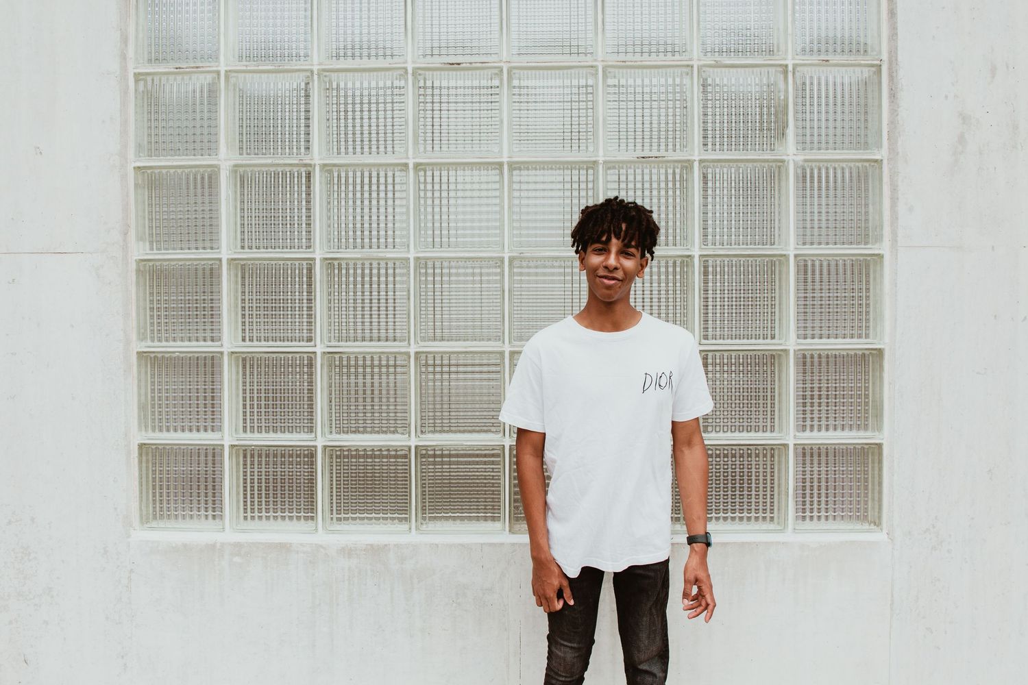 Young man in white t-shirt and sunglasses posing confidently against a glass block wall outdoors.