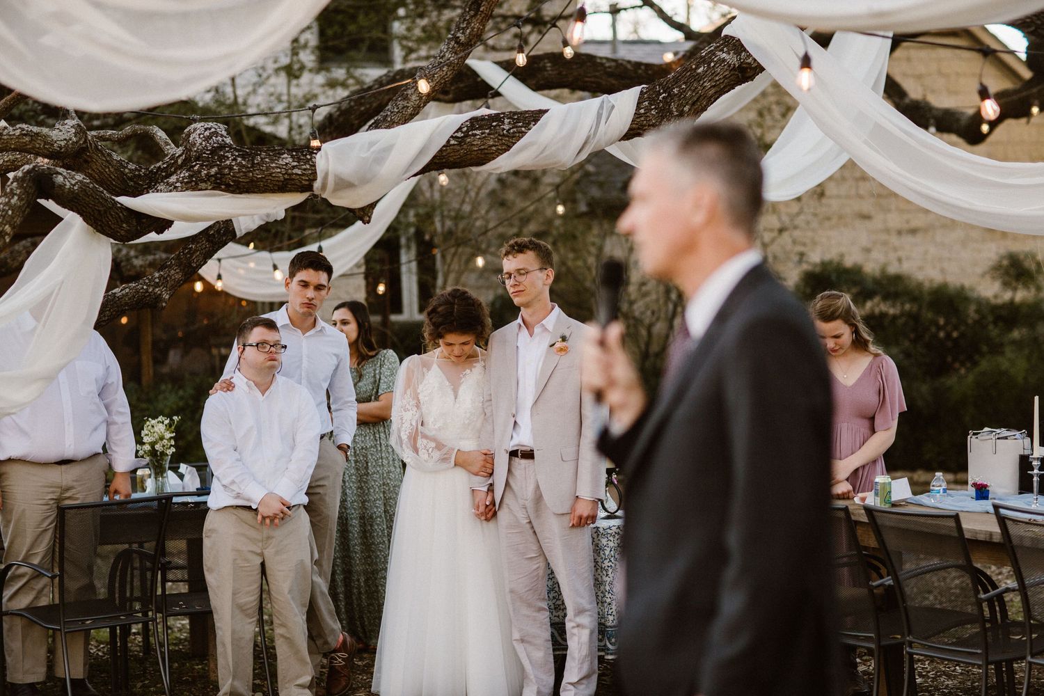 Wedding guests watch as a couple listens to a speech at an elegant outdoor reception.