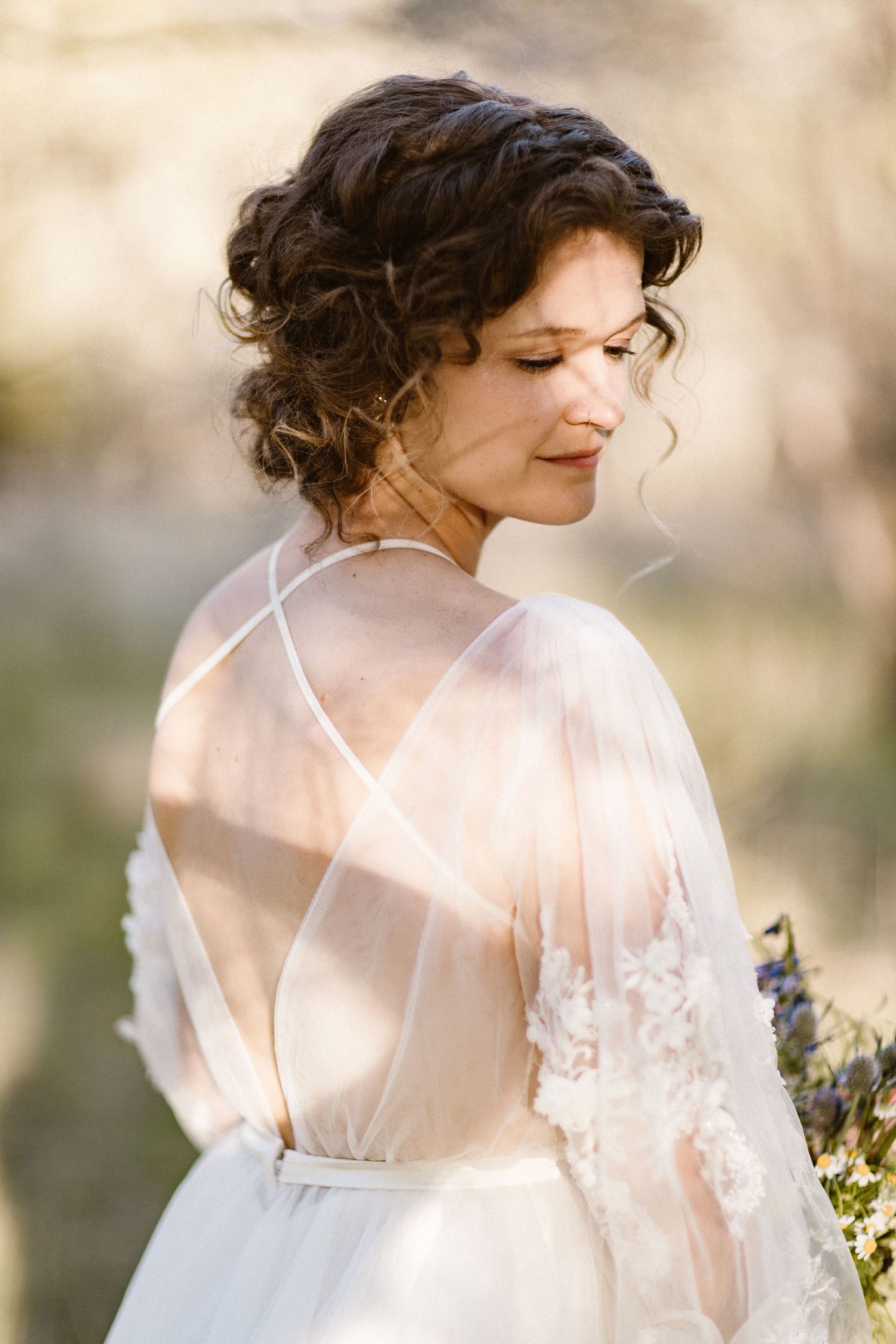 Elegant bride in sheer lace wedding dress with open back standing in golden sunlight outdoors.
