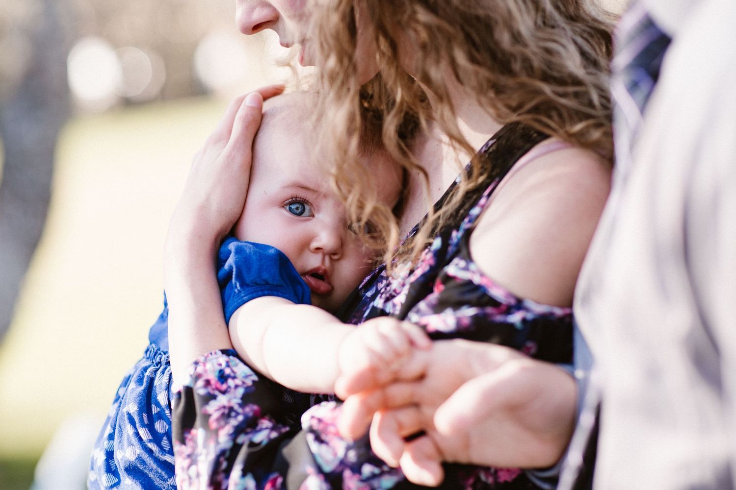 Mother in floral dress tenderly holds sleeping baby in blue outfit during outdoor wedding celebration.