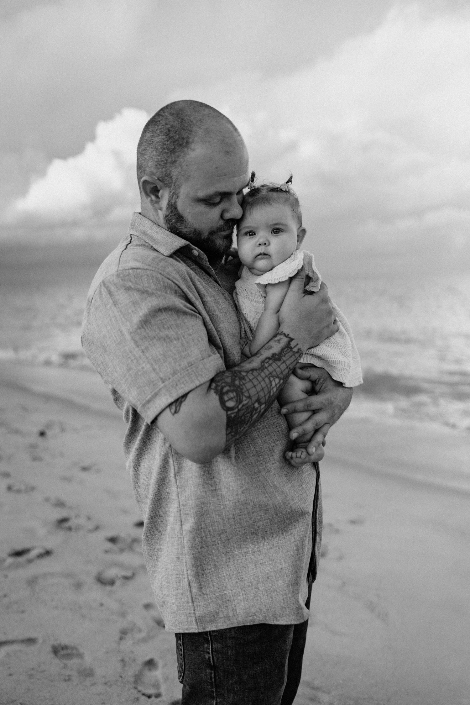 Black and white photo of tattooed father tenderly holding infant baby on a beach.