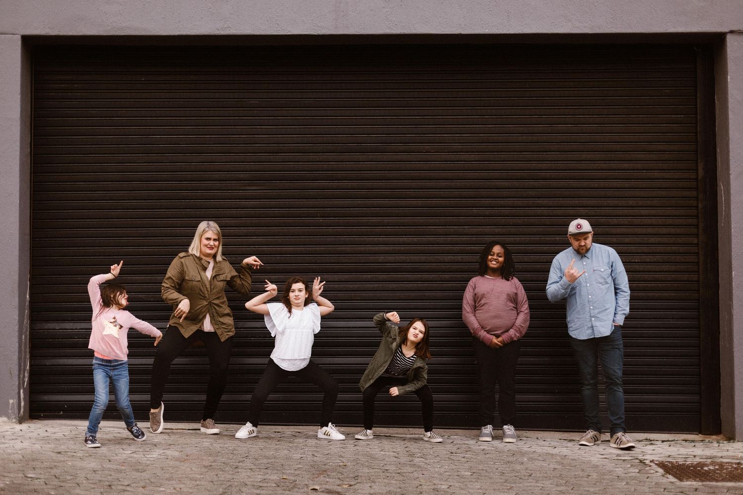 Fun family of six striking playful poses in front of a large dark brown garage door.