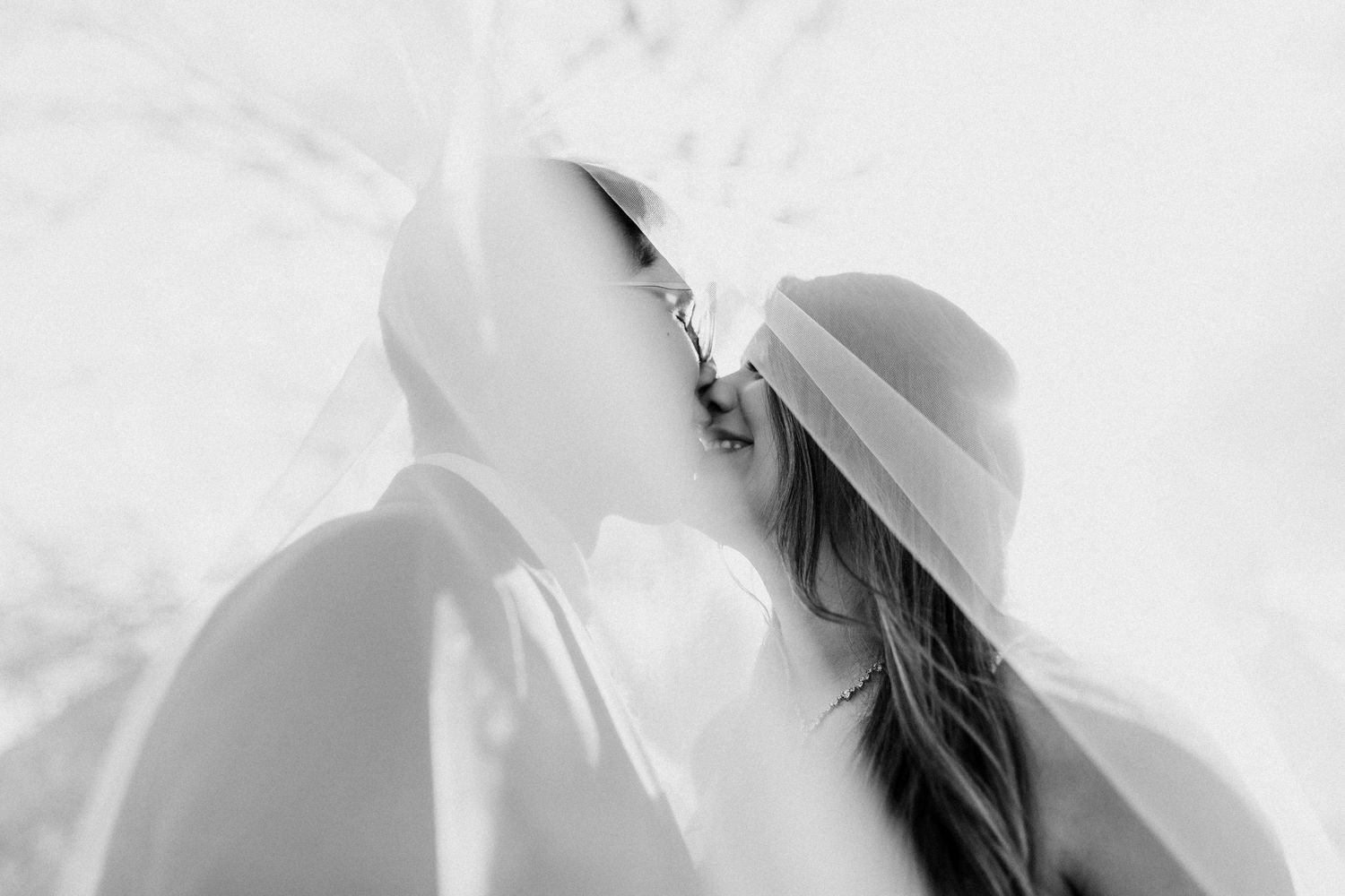 Romantic black and white photo of bride and groom kissing under flowing wedding veil outdoors.