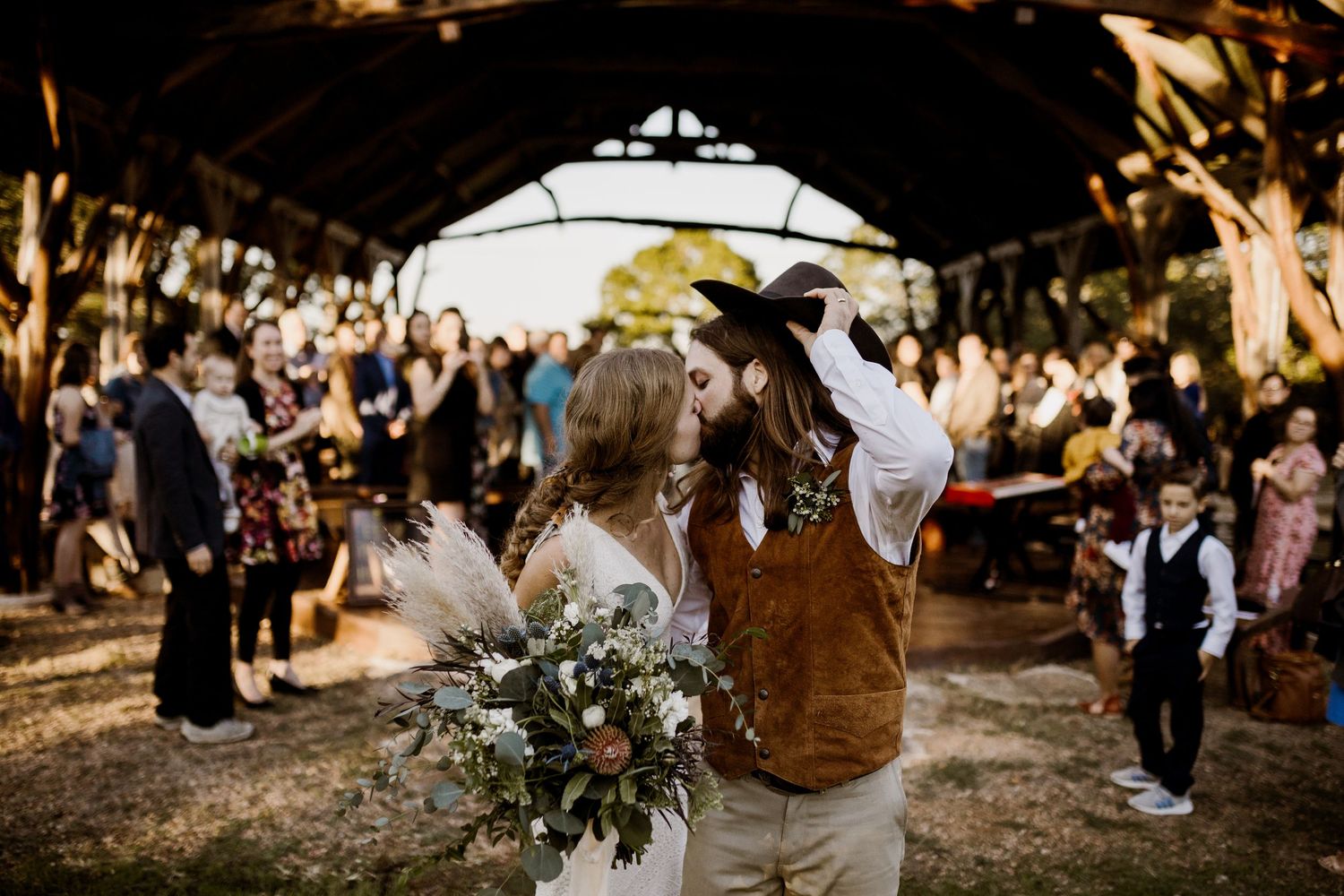 Couple sharing first kiss at rustic outdoor wedding ceremony with guests watching.
