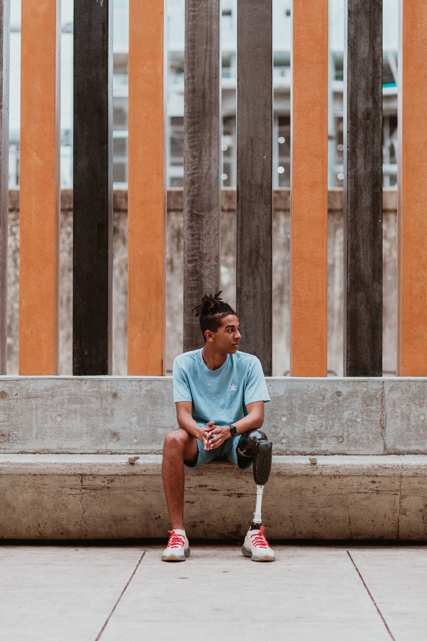 Young man with a prosthetic leg sitting on a concrete bench against a wooden fence in an urban setting.