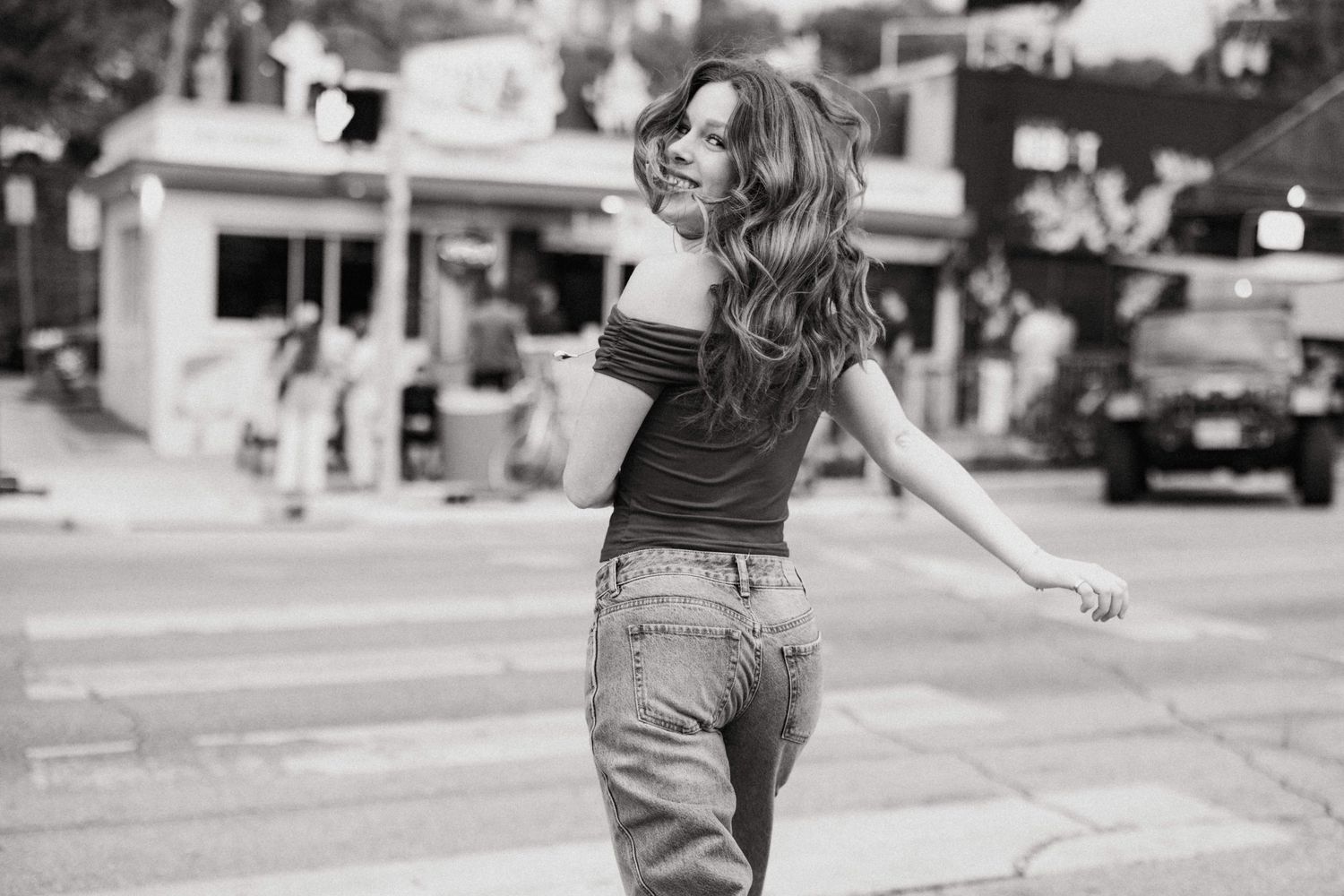 Black and white photo of woman in jeans twirling on a city street crosswalk.