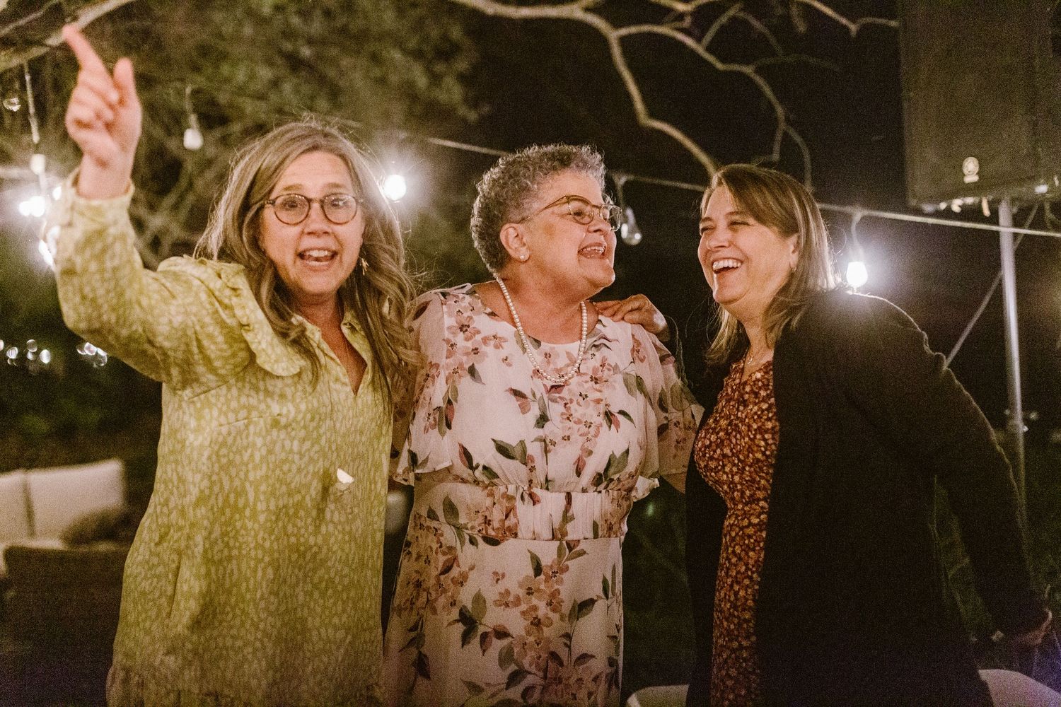 Three women laughing and pointing at string lights during an outdoor evening celebration.