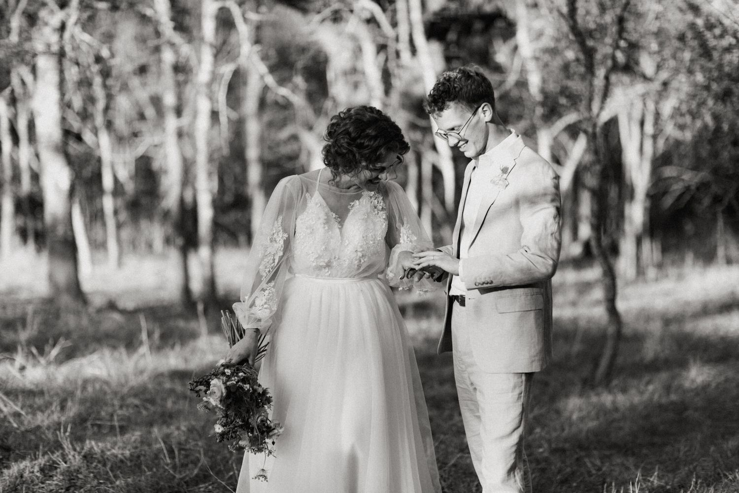 Black and white photo of bride and groom exchanging rings in a forest clearing during outdoor wedding ceremony.