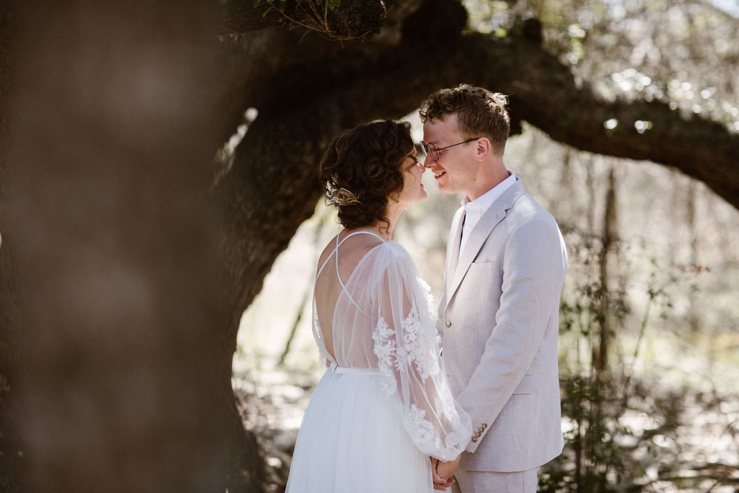 Bride and groom sharing an intimate moment holding hands under a sunlit oak tree.