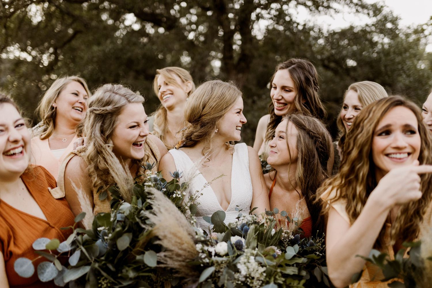 Joyful bride surrounded by laughing bridesmaids in rust and orange dresses holding greenery bouquets.
