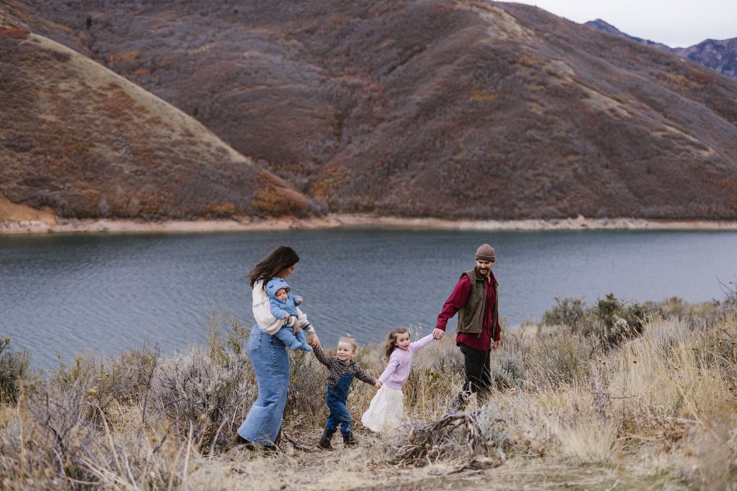Family of four holding hands by a scenic mountain lake surrounded by dry brush in autumn.