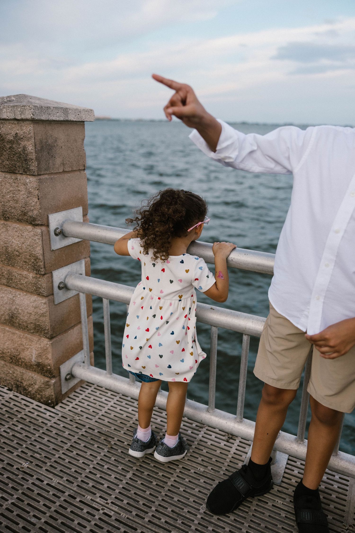 Young curly-haired girl in heart-print dress looking at ocean through pier railing as father points into distance.